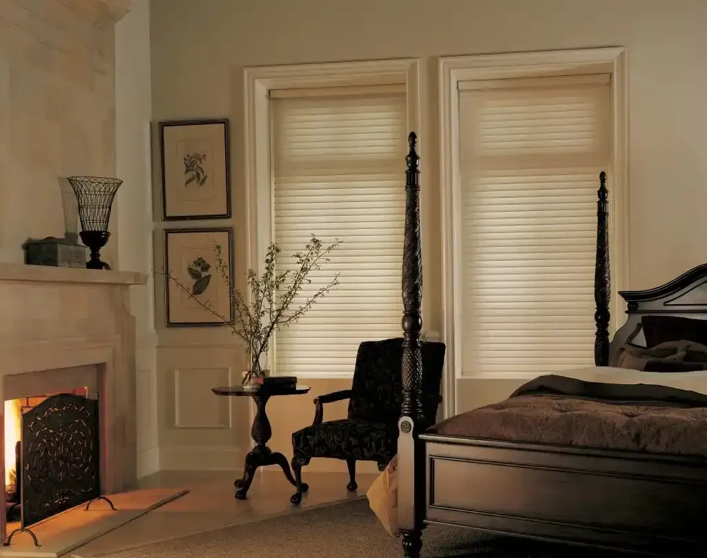 Bedroom with fireplace, dark wood bed and chair, two windows with blinds, and artwork.