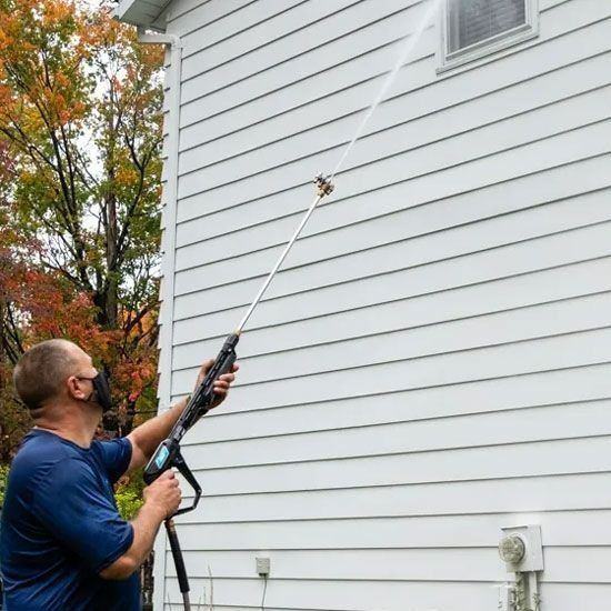 Man pressure washing a white house exterior, aiming at a window. He wears a mask.