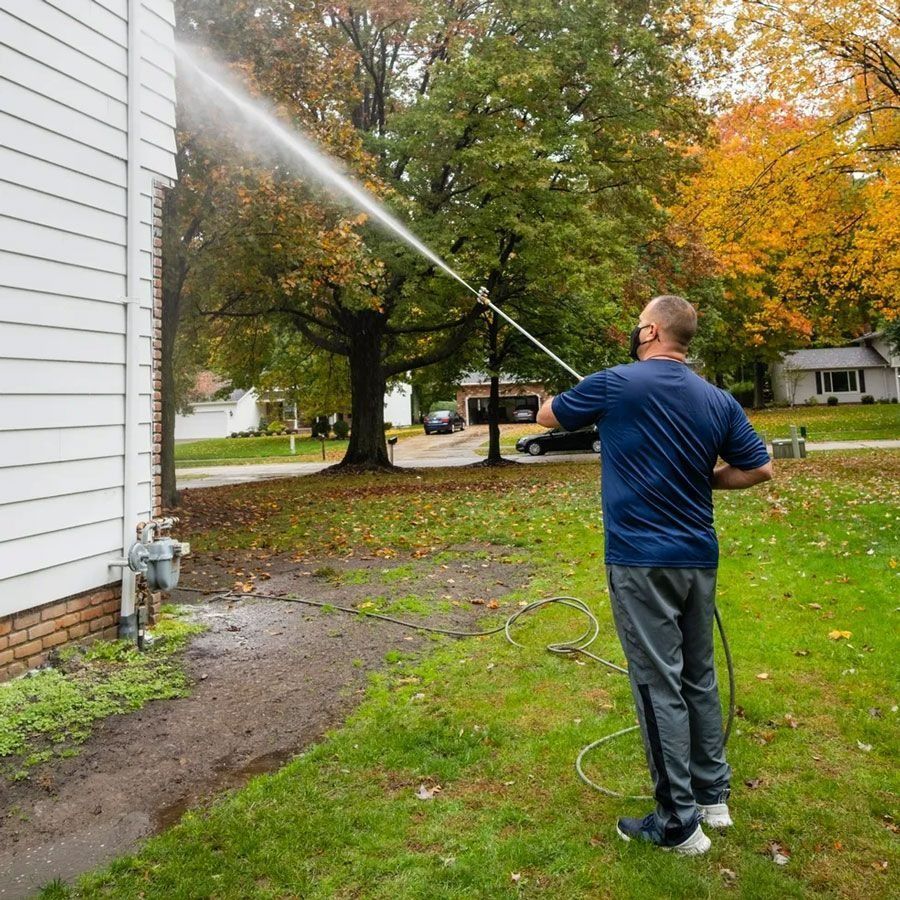 Man pressure washing the side of a white house with a long nozzle. Autumn trees in the background.