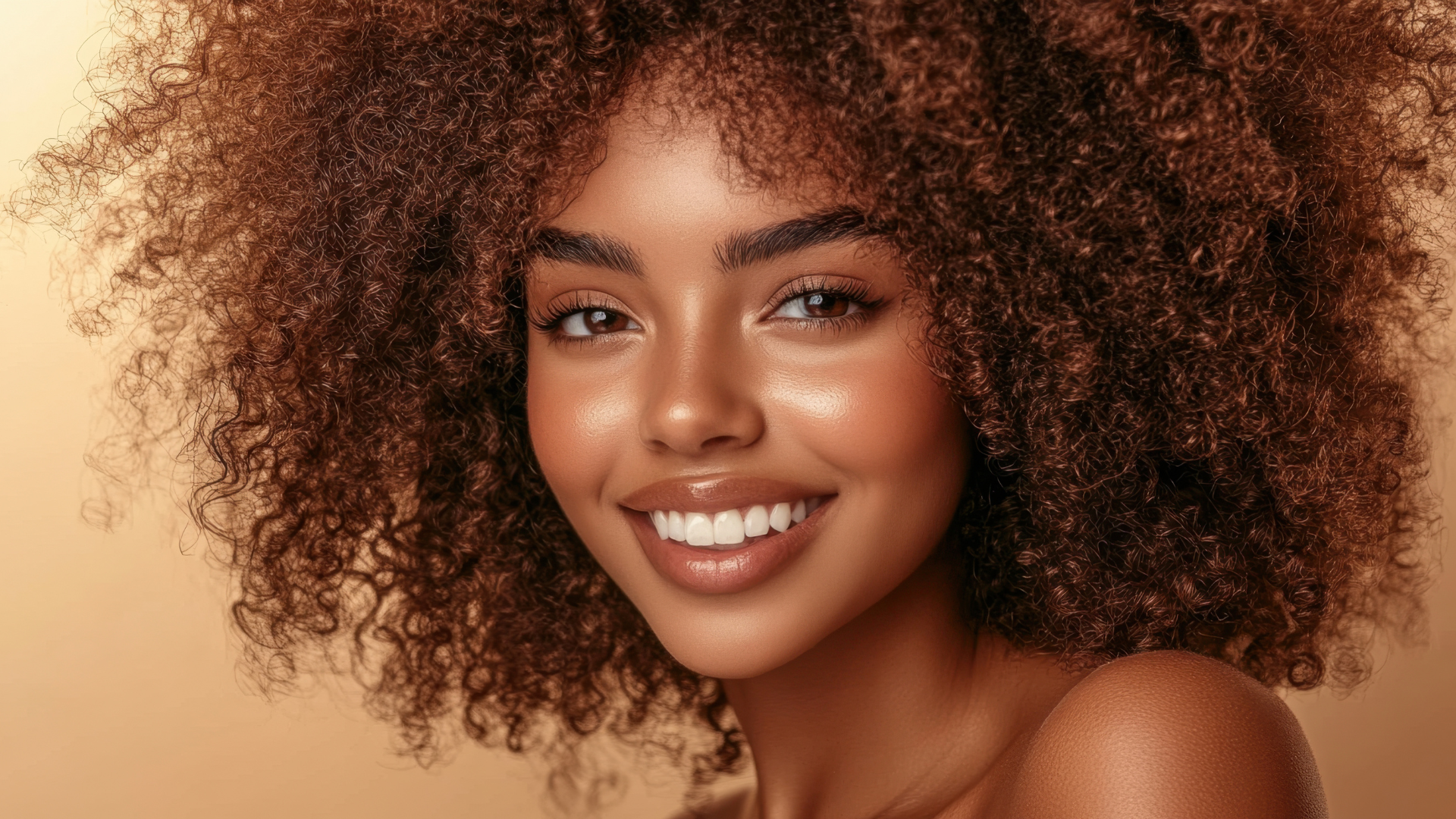 Frau mit lockigem braunem Haar und strahlender Haut lächelt vor einem hellbraunen Hintergrund.