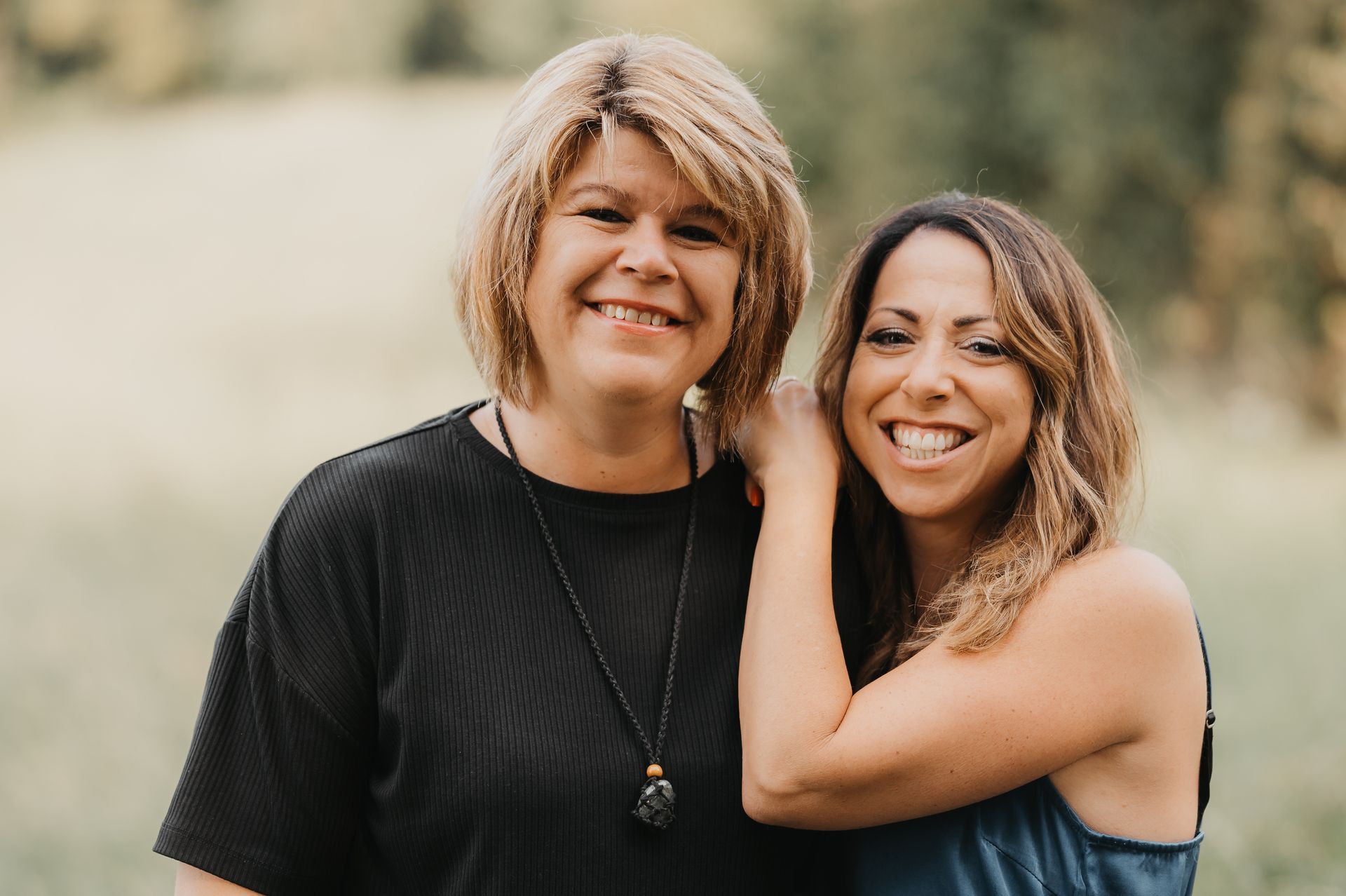 Zwei lächelnde Frauen, die eine mit kurzen blonden Haaren und einem schwarzen Oberteil, die andere mit langen braunen Haaren und einem blauen Oberteil, stehen im Freien.