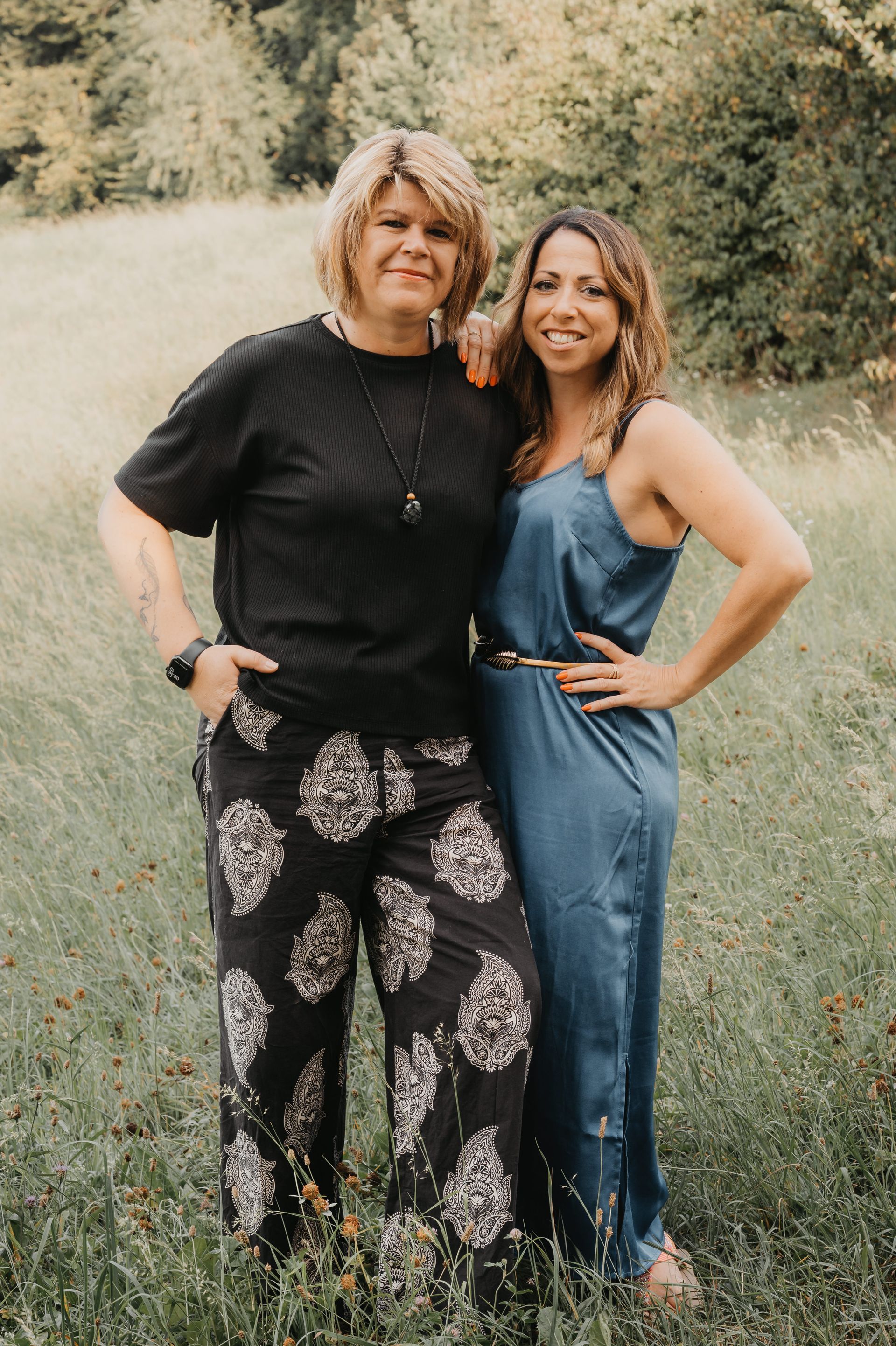 Zwei Frauen stehen auf einer Wiese. Die eine trägt ein schwarzes Oberteil und eine gemusterte Hose, die andere einen blauen Overall.