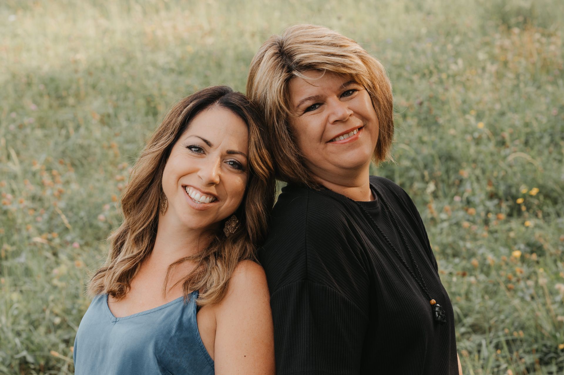 Zwei Frauen lächeln, Rücken an Rücken in einer grünen Wiese. Die Frau links trägt Blau, die andere Schwarz.