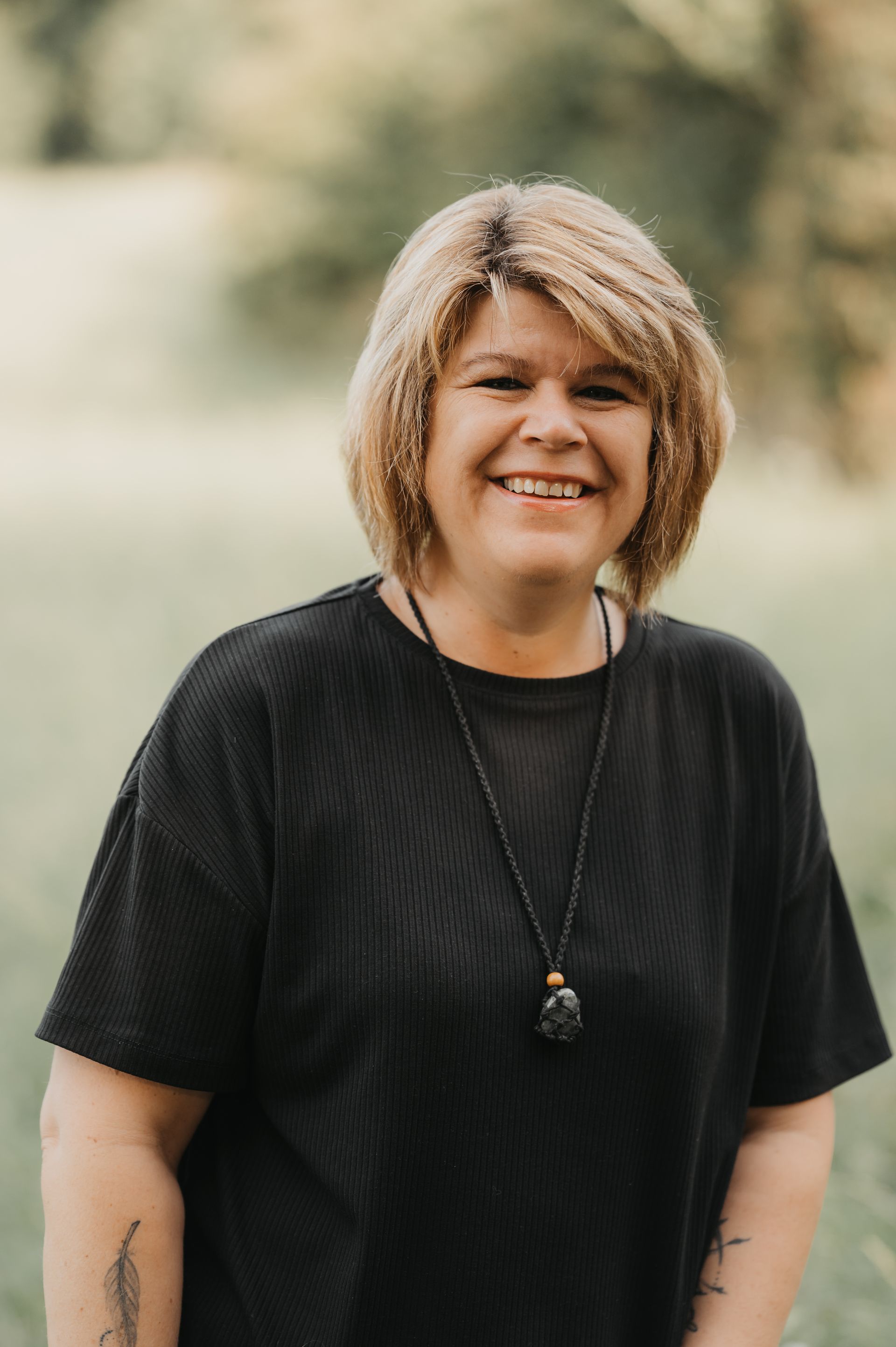 Eine lächelnde Frau mit blonden Haaren, die ein schwarzes Hemd und eine schwarze Halskette trägt, steht im Freien.