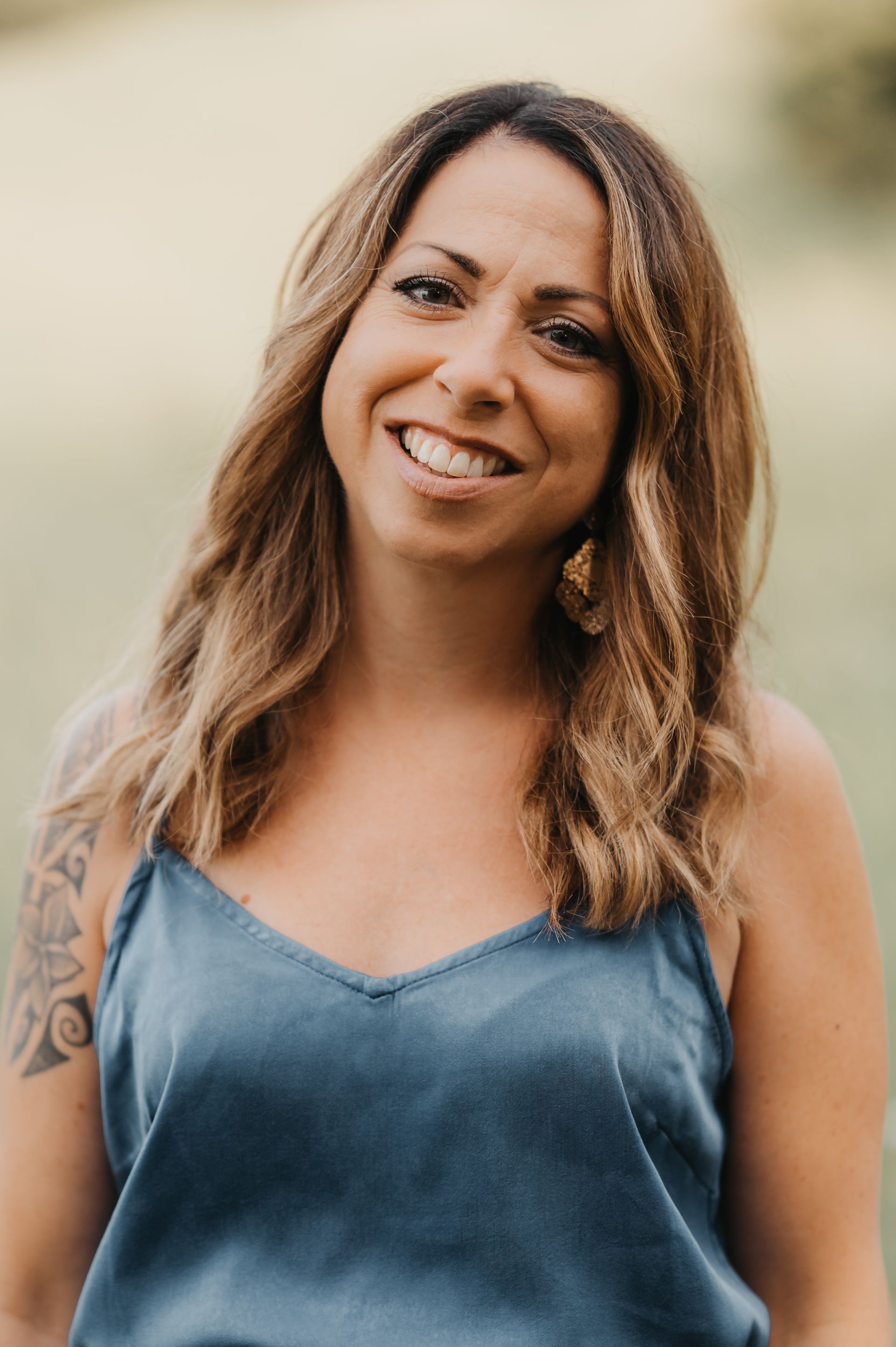 Frau mit langen, gewellten braunen Haaren, lächelnd, trägt ein blaues Tanktop, im Freien.