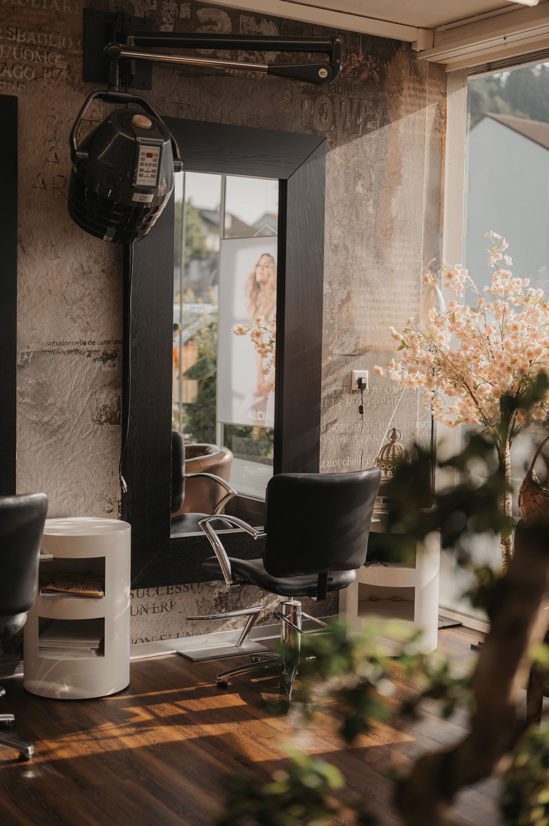 Saloninterieur mit großem Spiegel, Friseurstuhl und Haartrockner mit Haube. Sonnenlicht strömt durch ein Fenster herein.