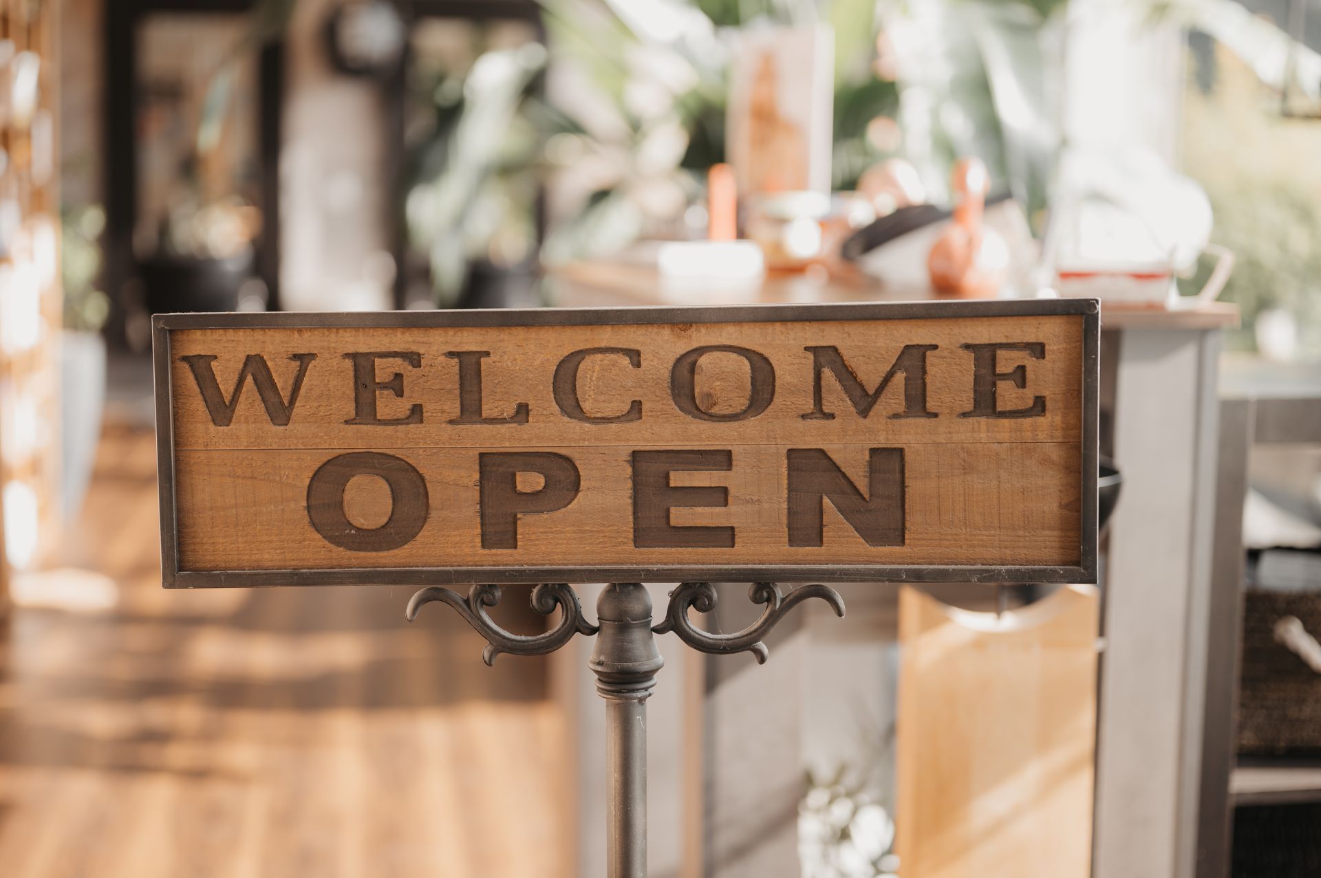 Ein Schild mit der Aufschrift „Willkommen geöffnet“ in einem Geschäft, braunes Schild mit dekorativem Metallständer, verschwommener Innenraumhintergrund.