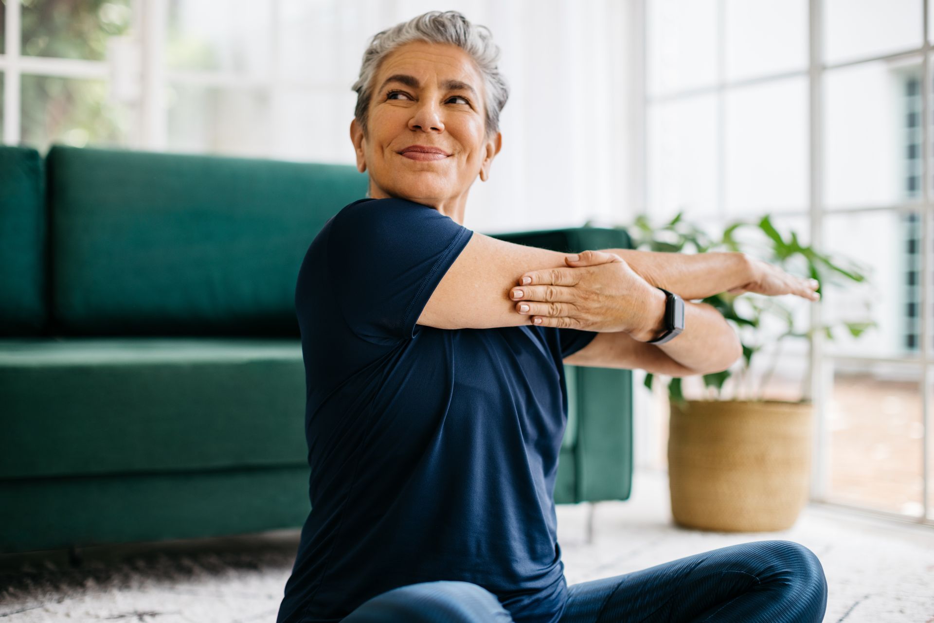 A healthy senior woman doing a cross-arm stretch while seated in her space.