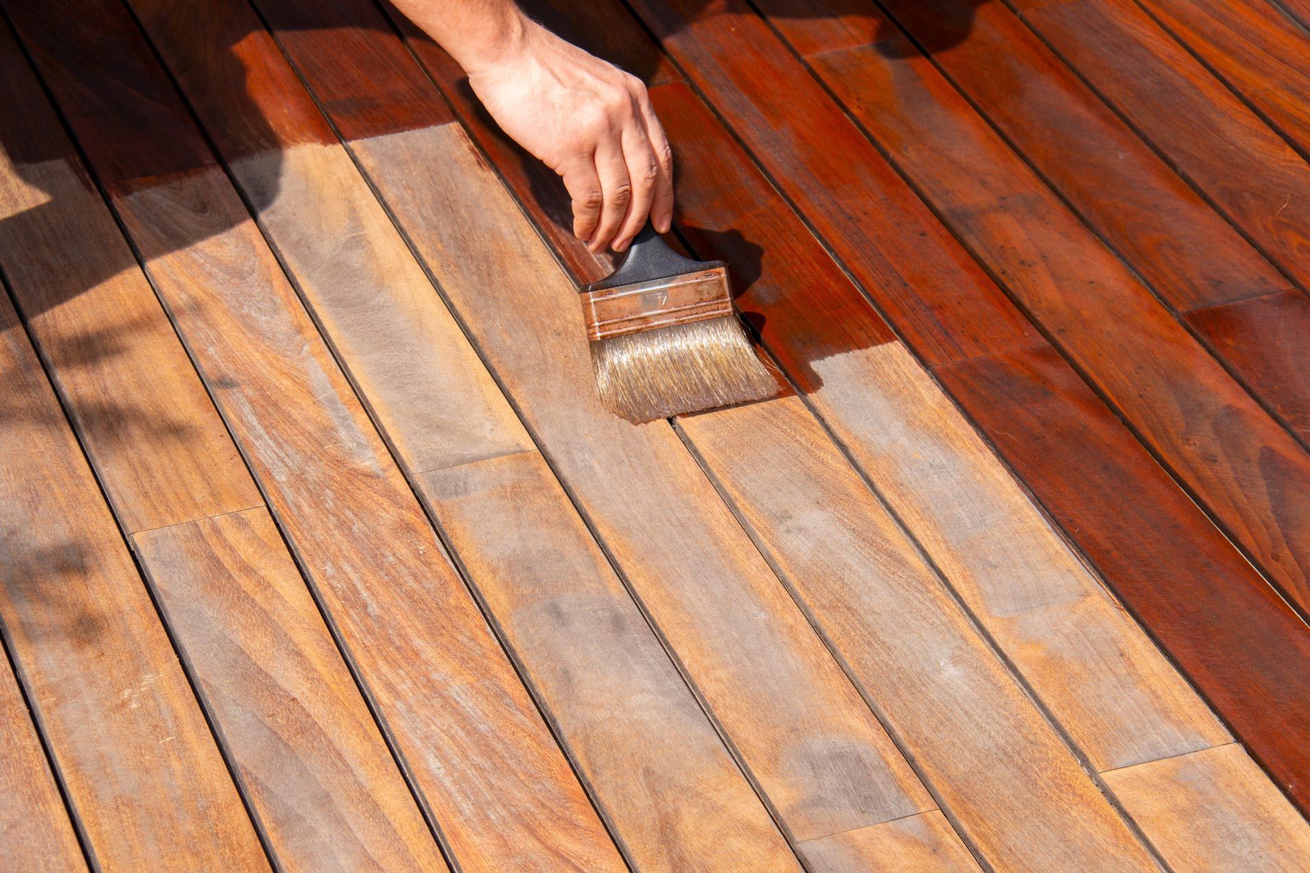 Person brushing wood stain on a wooden deck, changing the color from light to dark brown.