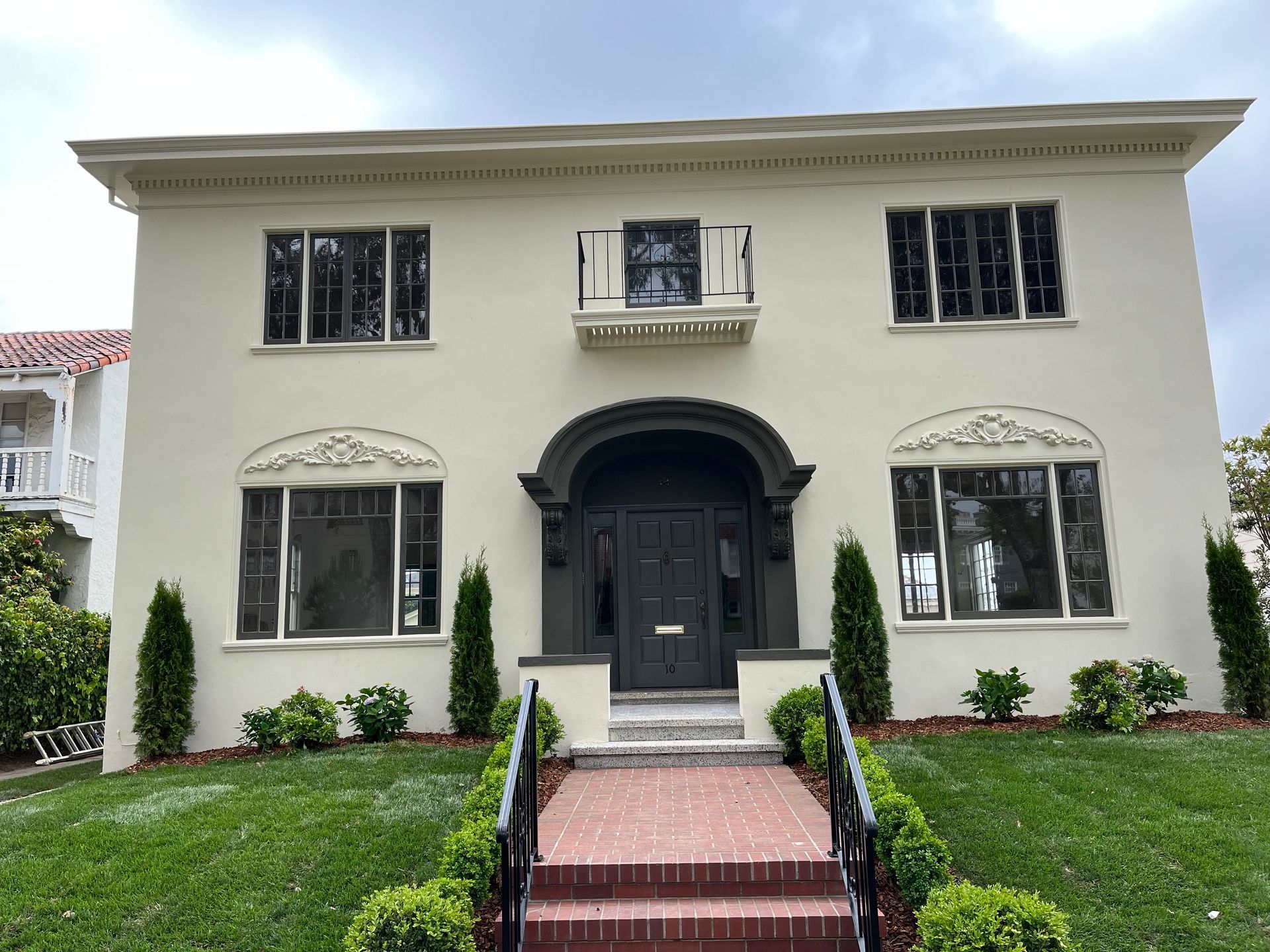 Man painting exterior window trim on a beige house with white shutters and blue sky.