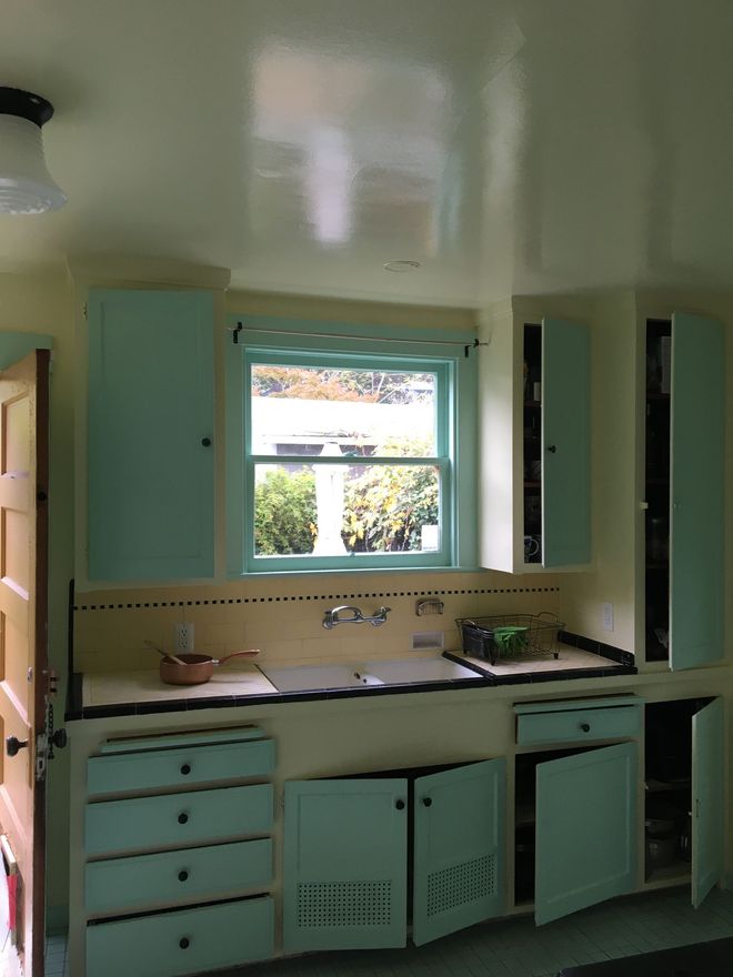 A person in blue overalls painting a white border on a teal wall in a room.