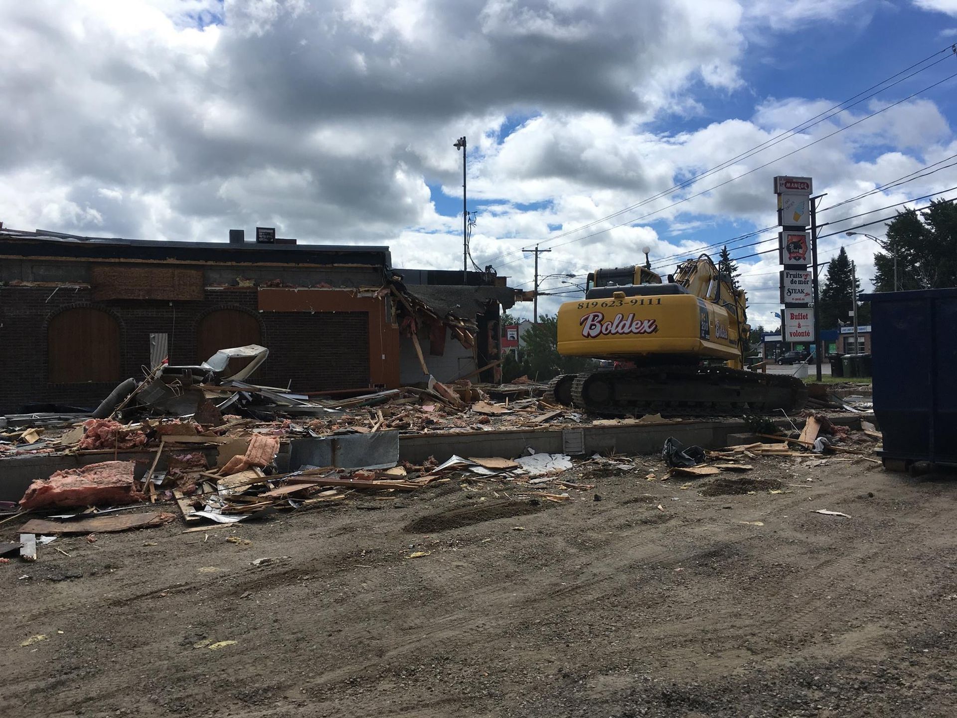 Démolition d'un chantier à l'aide d'une excavatrice sous un ciel nuageux. Débris et fondations apparentes.