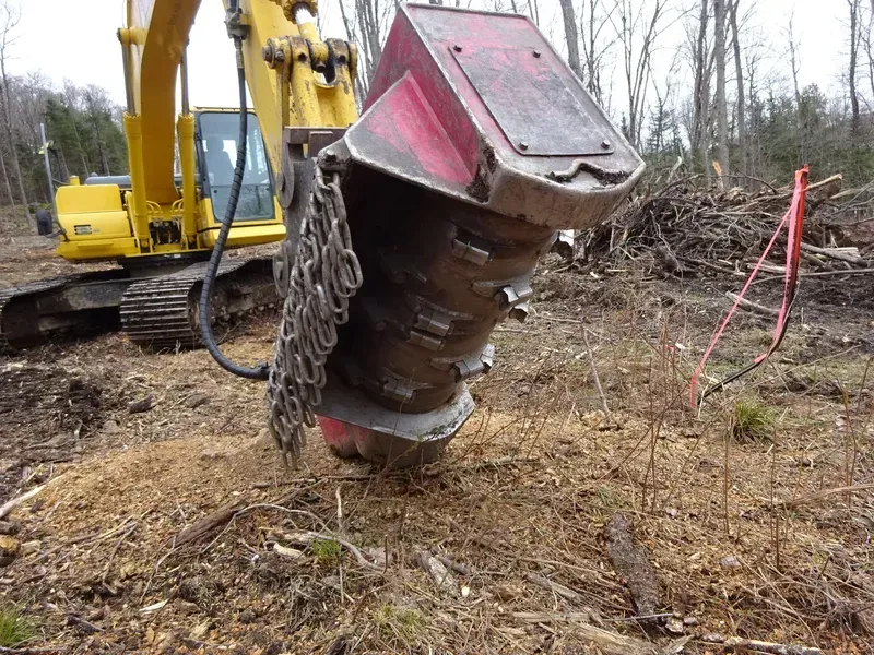 Une pelleteuse jaune équipée d'une débroussailleuse rouge broie des copeaux de bois dans une clairière.