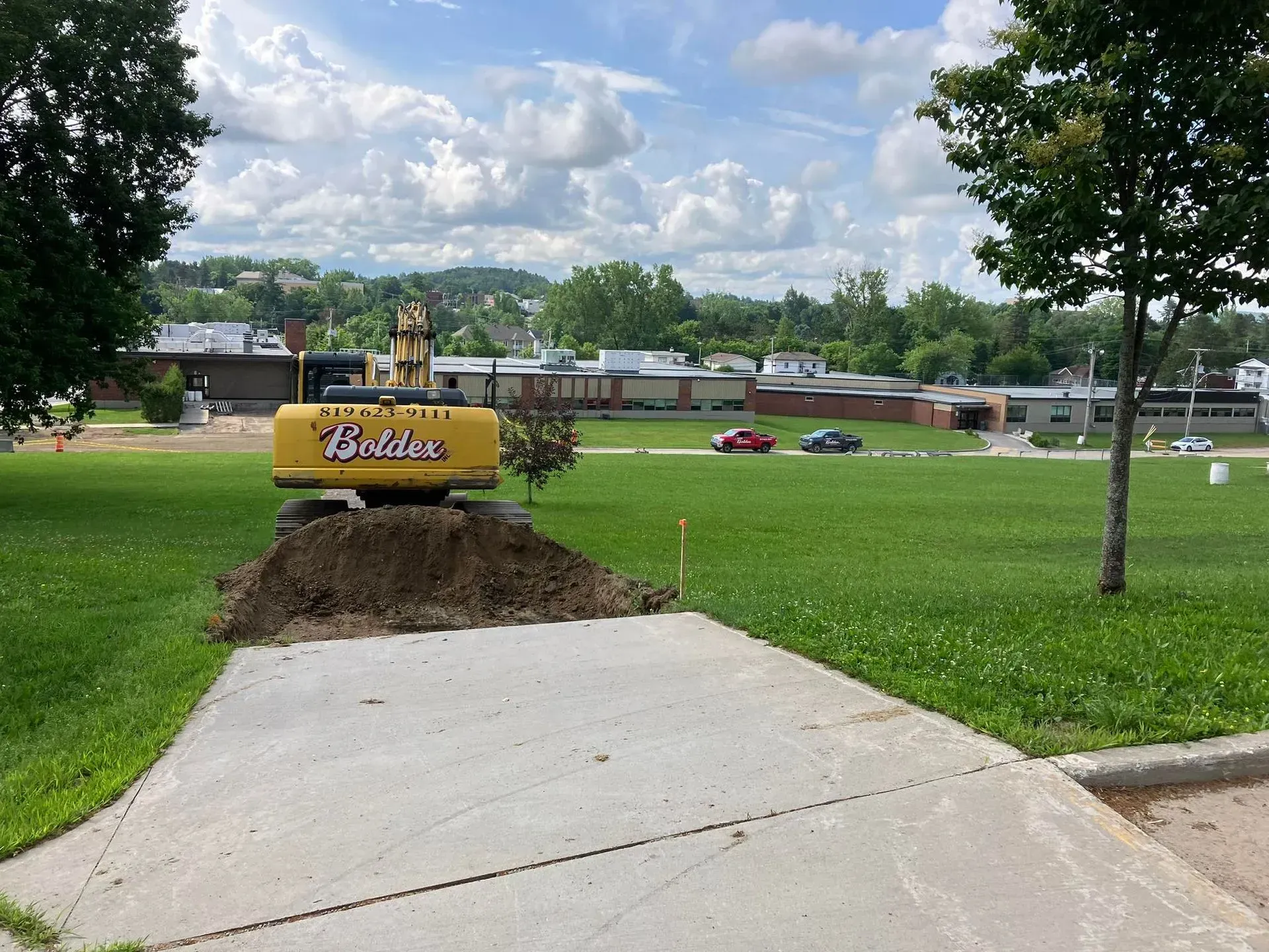 Une pelleteuse jaune creuse la terre sur l'herbe à côté d'une allée en béton, avec des bâtiments en arrière-plan.