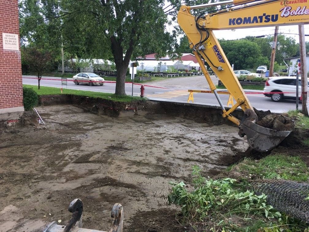Une pelleteuse jaune creuse la terre au bord d'une rue bordée de voitures, d'herbe et d'un bâtiment en briques.