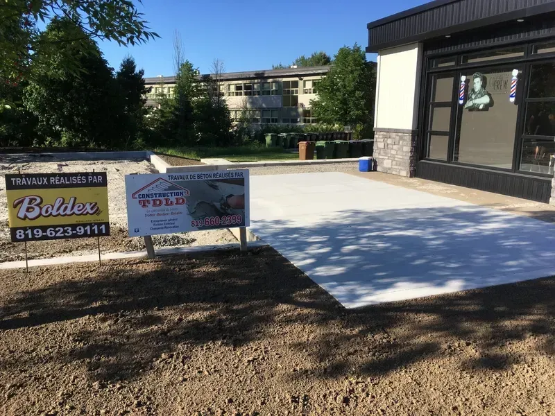 Terrasse en béton fraîchement coulée jouxtant un bâtiment commercial, avec des panneaux indiquant une entreprise de construction.