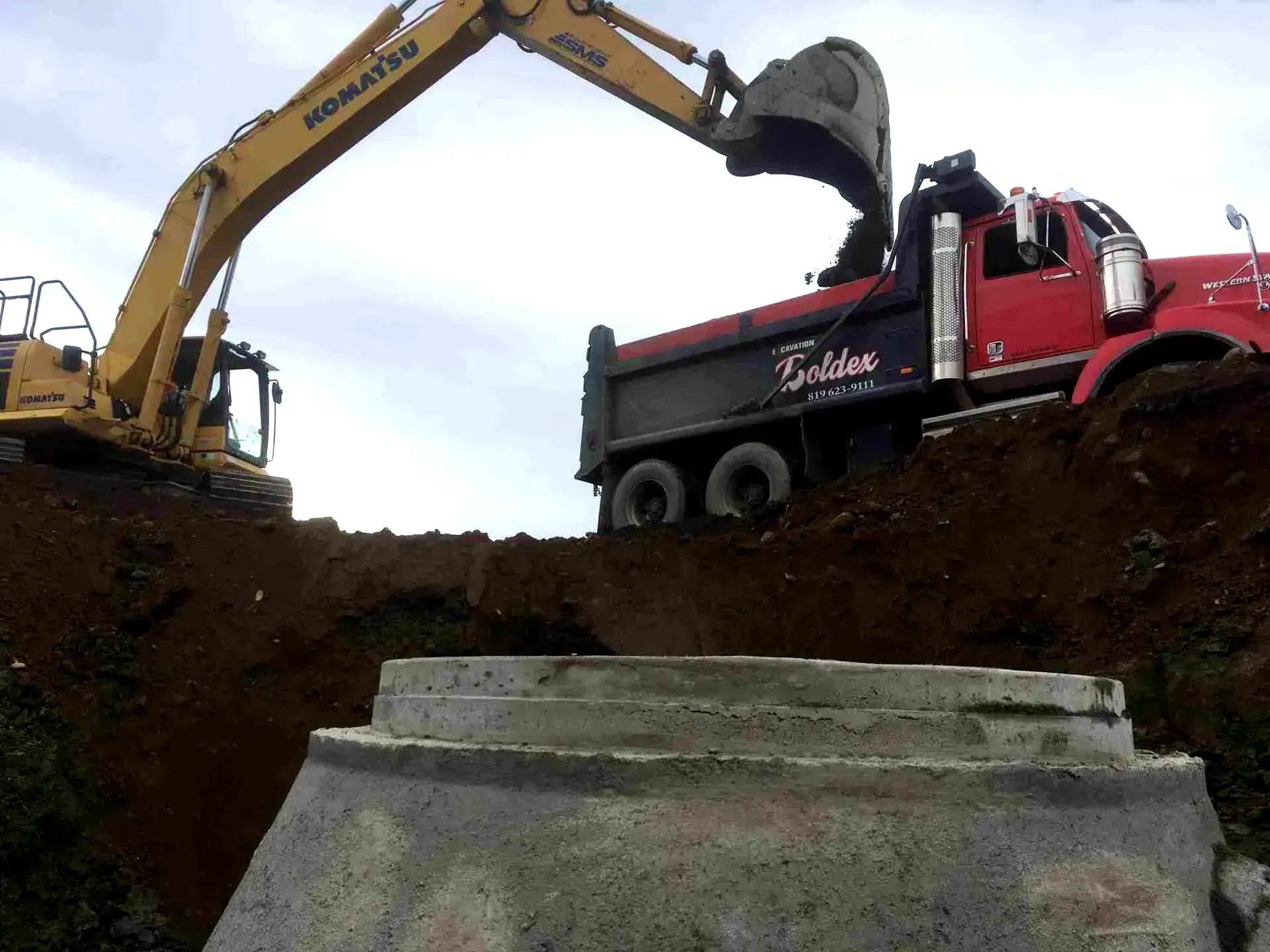 Une pelleteuse jaune charge de la terre dans un camion-benne rouge, à côté d'une structure en béton. Ciel nuageux.