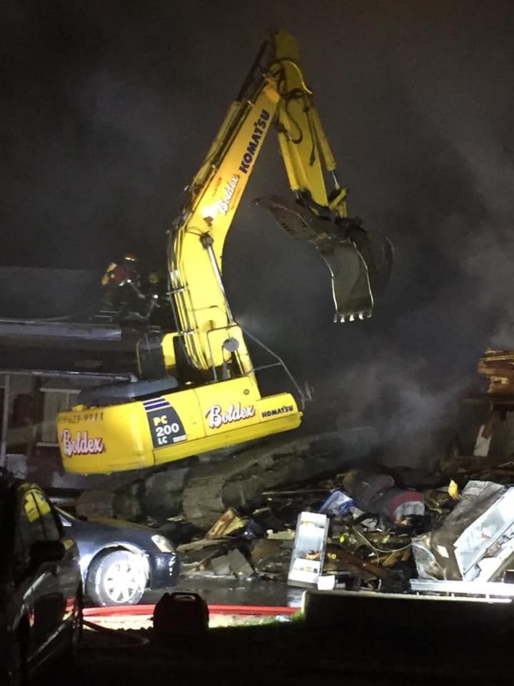 Une pelle Komatsu jaune démolit un bâtiment de nuit. Débris et fumée visibles.