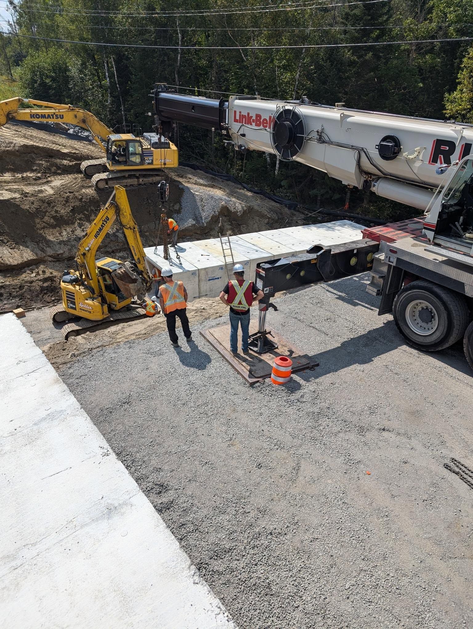 Chantier de construction avec des ouvriers, des excavatrices et une grue installant des blocs de béton.