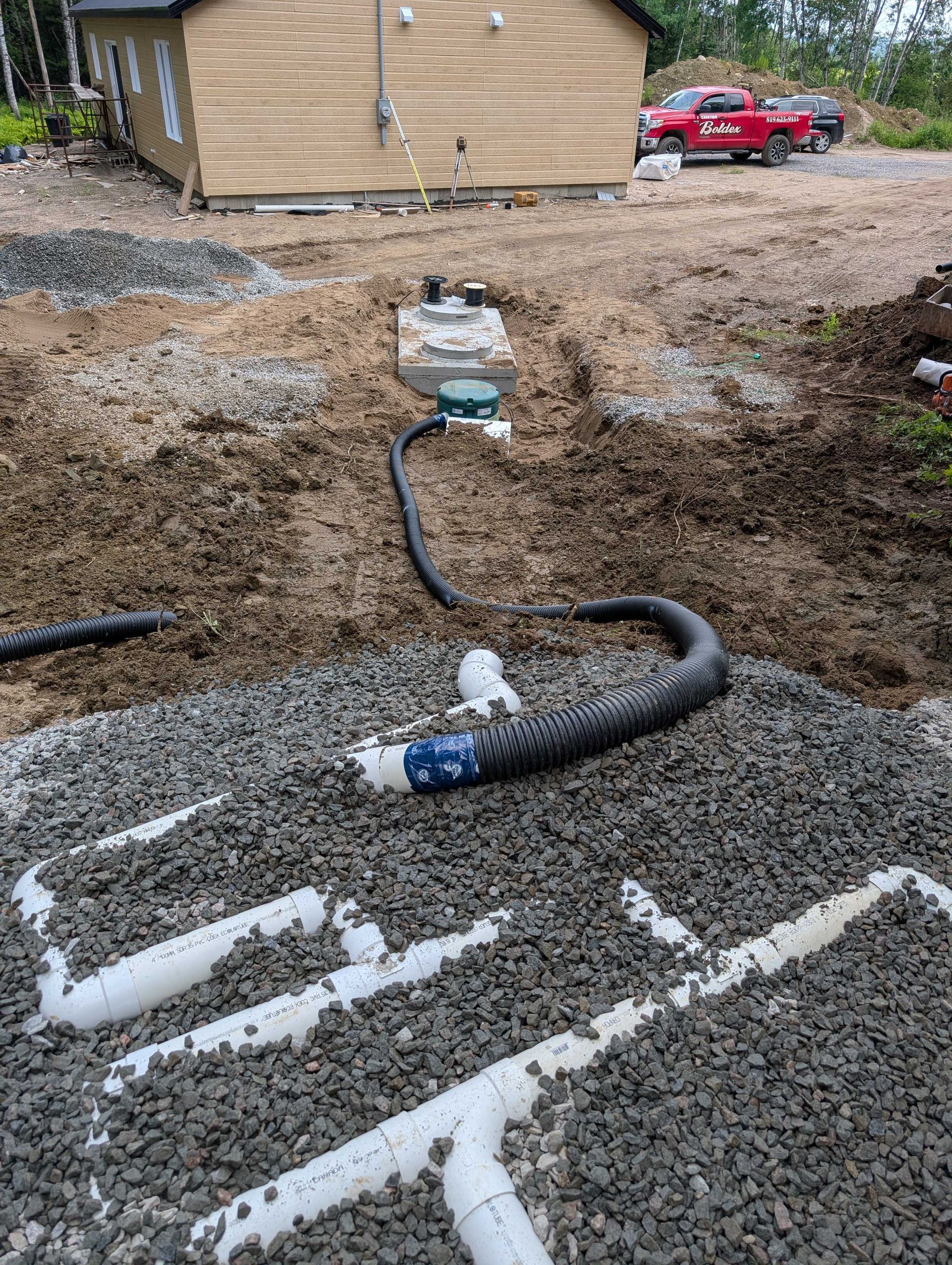 Installation d'une fosse septique : Excavation avec tuyaux et gravier, à proximité d'une maison et d'un camion.
