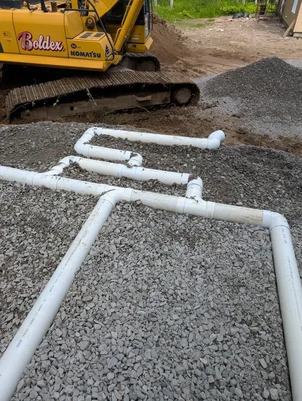 Des tuyaux en PVC blanc raccordés sur du gravier, près d'une excavatrice jaune sur un chantier de construction.