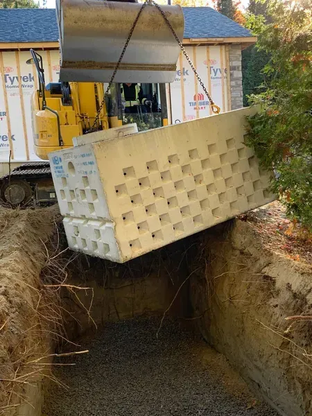 Une pelleteuse jaune descend une pile de blocs perforés de couleur claire dans une tranchée remplie de gravier ; un bâtiment se dresse en arrière-plan.