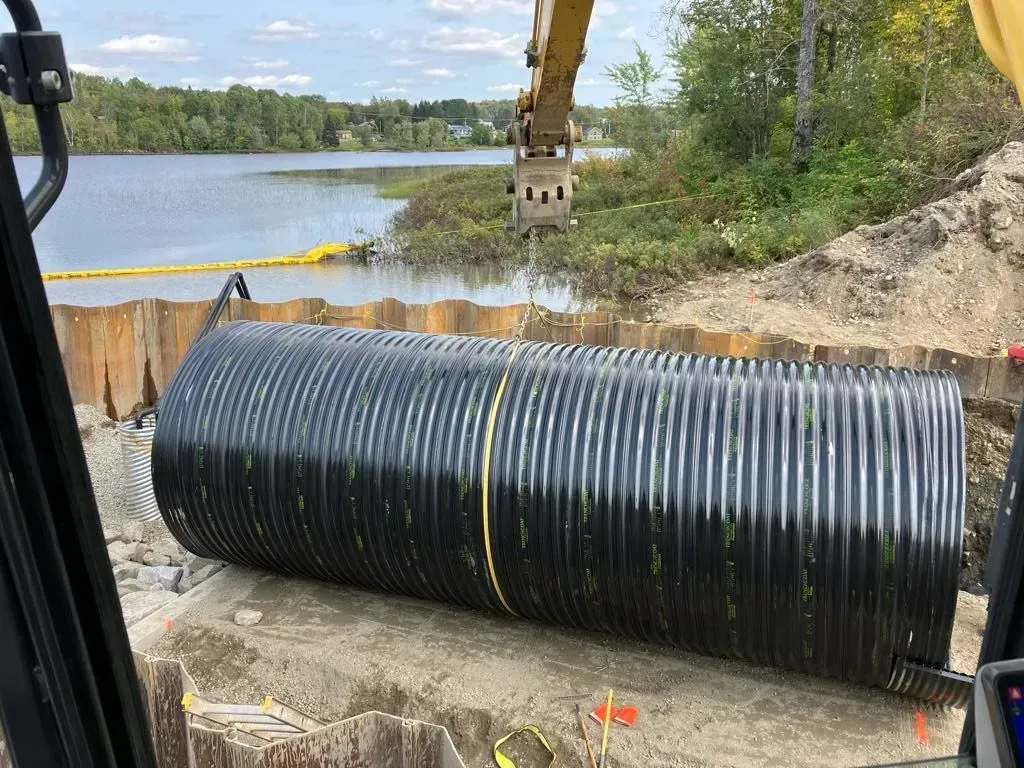 Un ponceau ondulé noir est installé par une excavatrice près d'un lac.