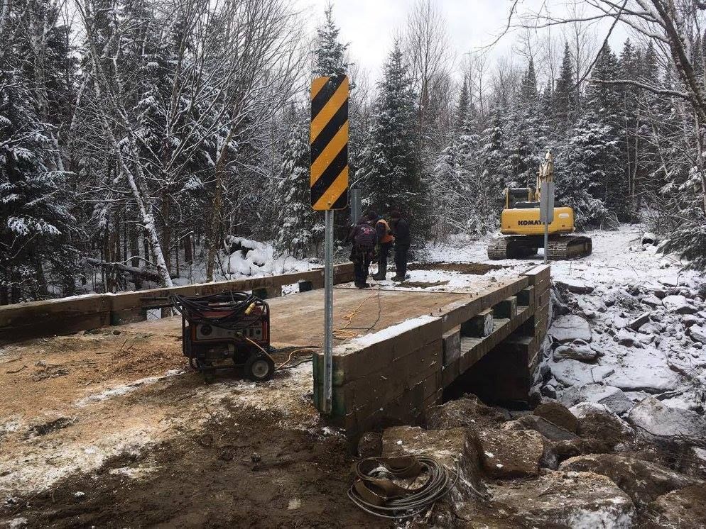 Construction d'un pont en forêt enneigée ; un générateur et une excavatrice sont présents.