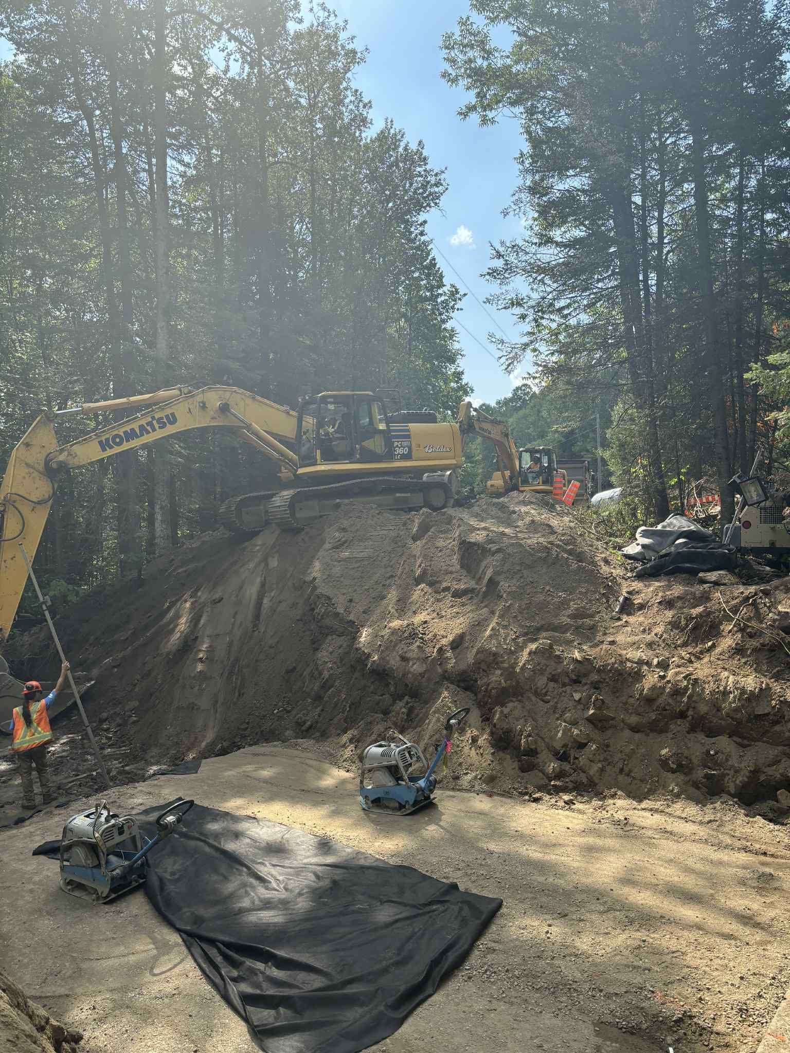 Pelle mécanique jaune sur un tas de terre, forêt en arrière-plan. Ouvriers du bâtiment sur le chantier, journée ensoleillée.