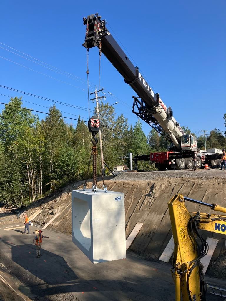 Une grue soulève une grande structure en béton. Chantier de construction avec ouvriers, excavatrice et lignes électriques.