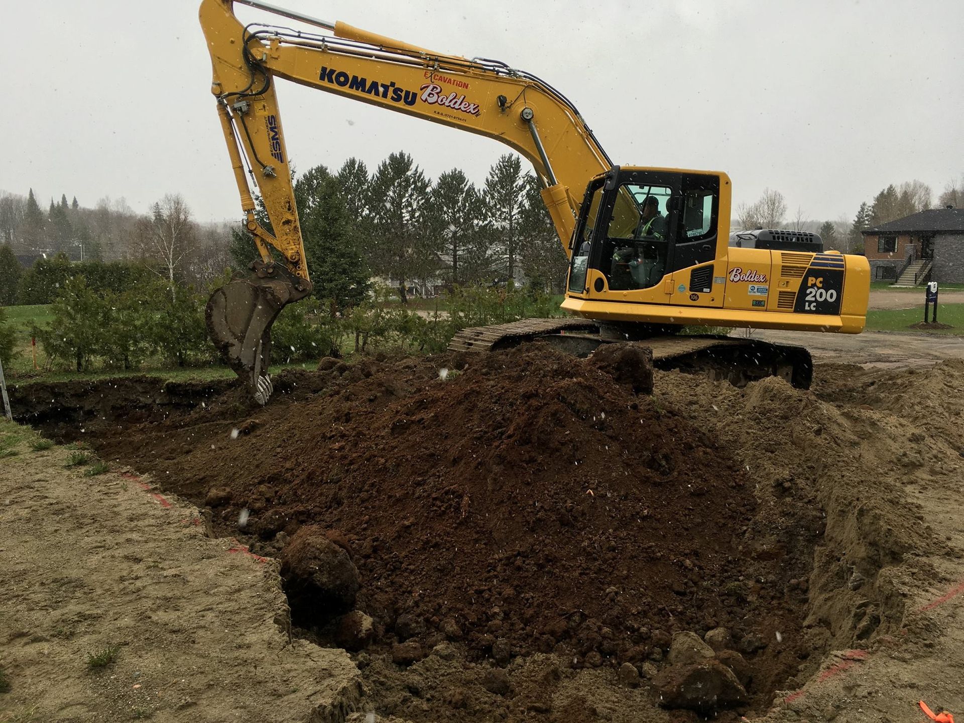 Une pelle mécanique Komatsu jaune creuse la terre sur un chantier de construction, avec un monticule de terre et des arbres en arrière-plan.