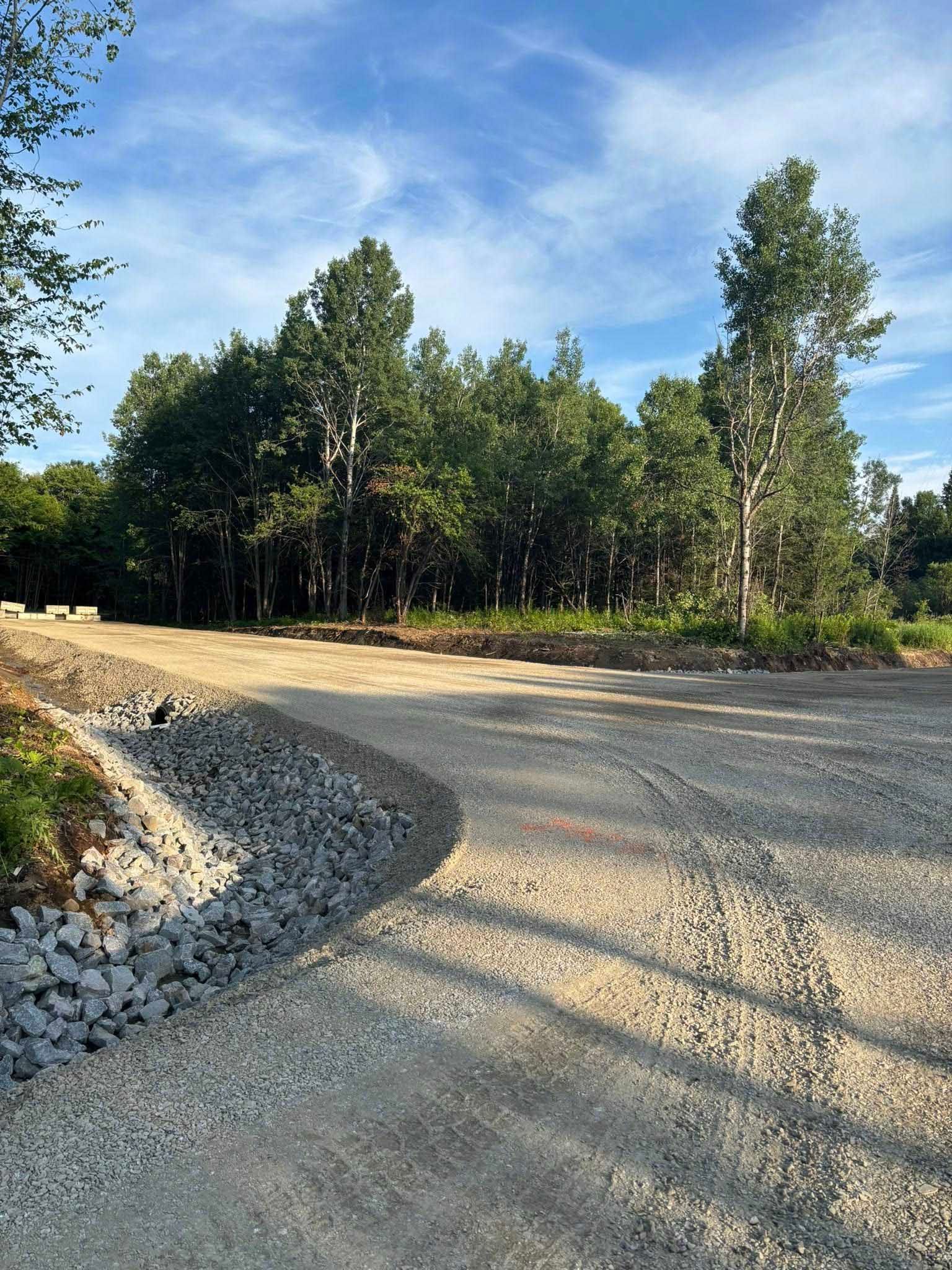 Un chemin de gravier serpente vers une lisière de forêt sous un ciel bleu, avec quelques rochers en bordure de route.