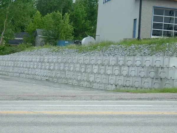Mur de soutènement en béton gris le long d'une route, retenant une zone en pente de gravier et d'herbe.