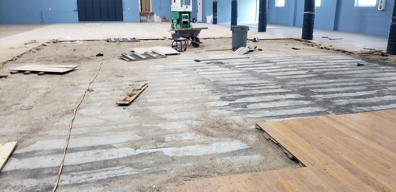 A construction site interior showing a partially removed floor, revealing concrete beneath wood flooring and debris.