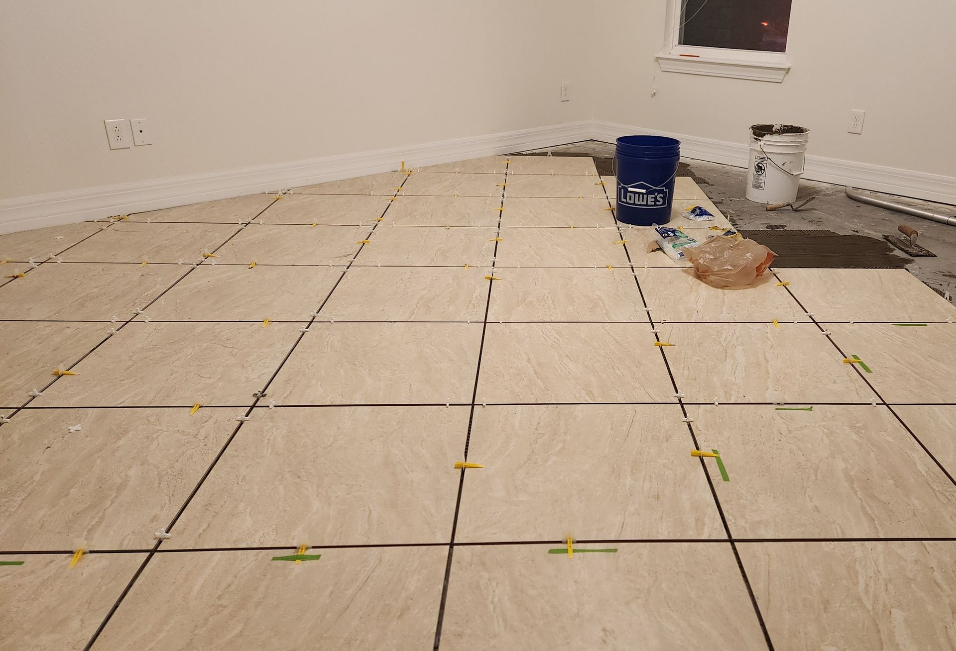 A light-colored tiled floor under construction with tile spacers, a blue bucket, and supplies in an unfinished room.