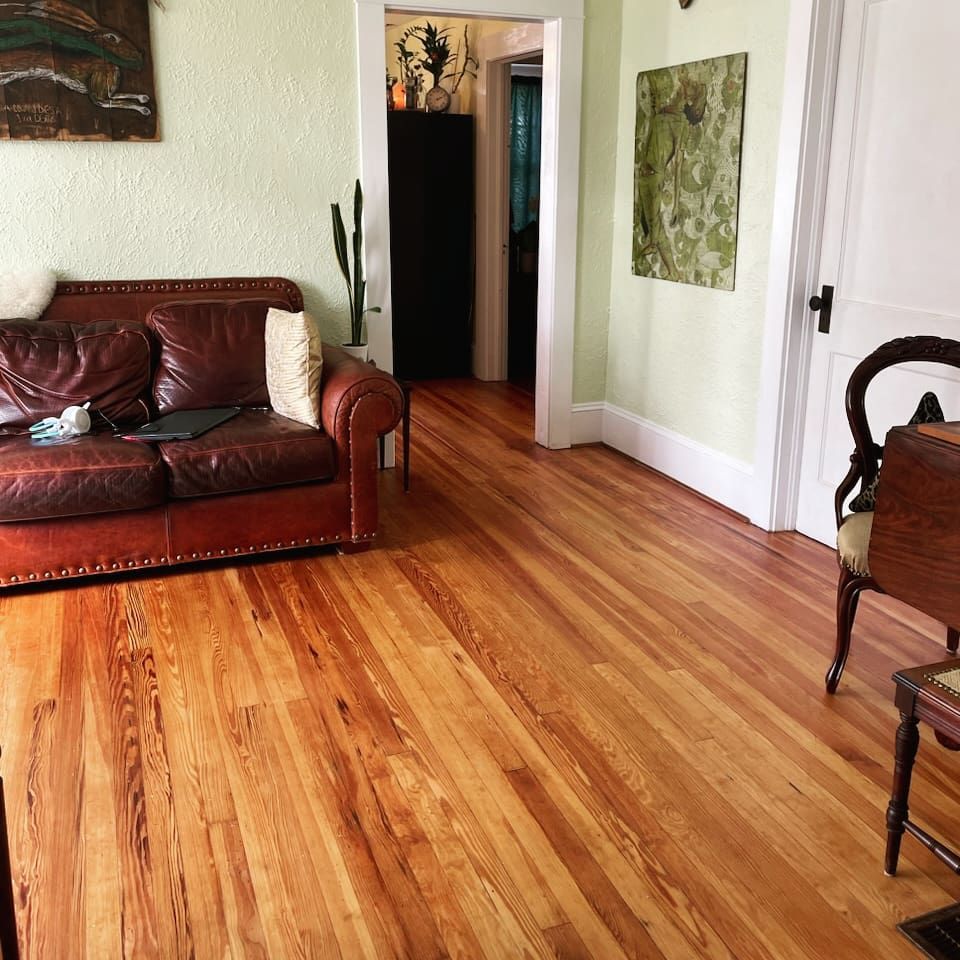 A leather sofa and wood chair sit on polished hardwood floors in a sunlit living room with pale green walls.