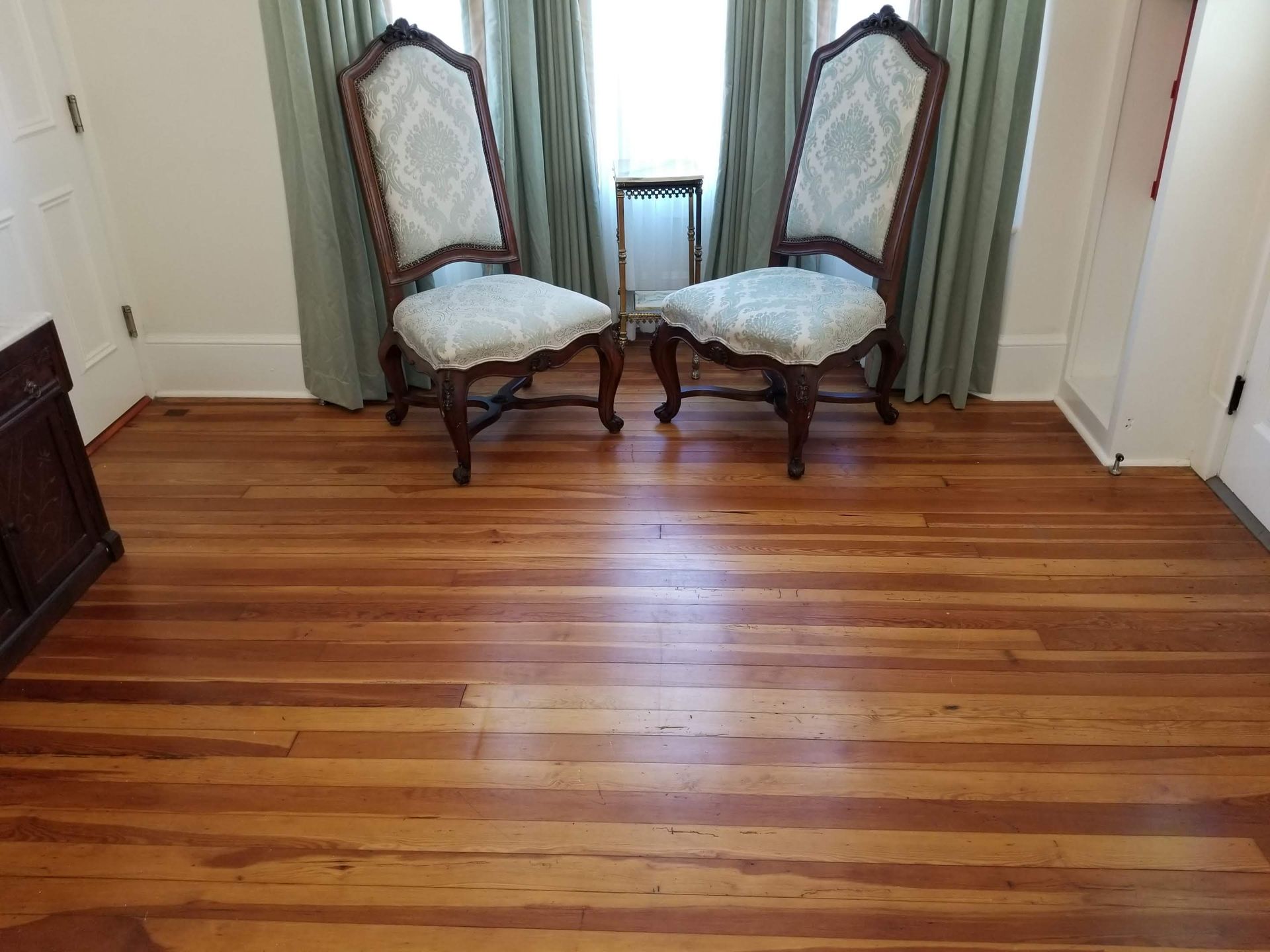 Two ornate, light-patterned chairs face forward in front of a window with sage green curtains, set on hardwood flooring.
