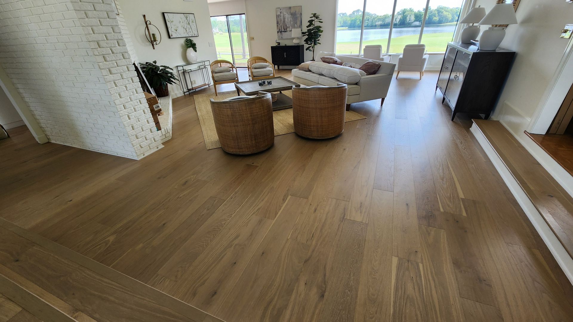 A sunlit, open-concept living room with light hardwood floors, woven armchairs, a sofa, and a white brick fireplace.