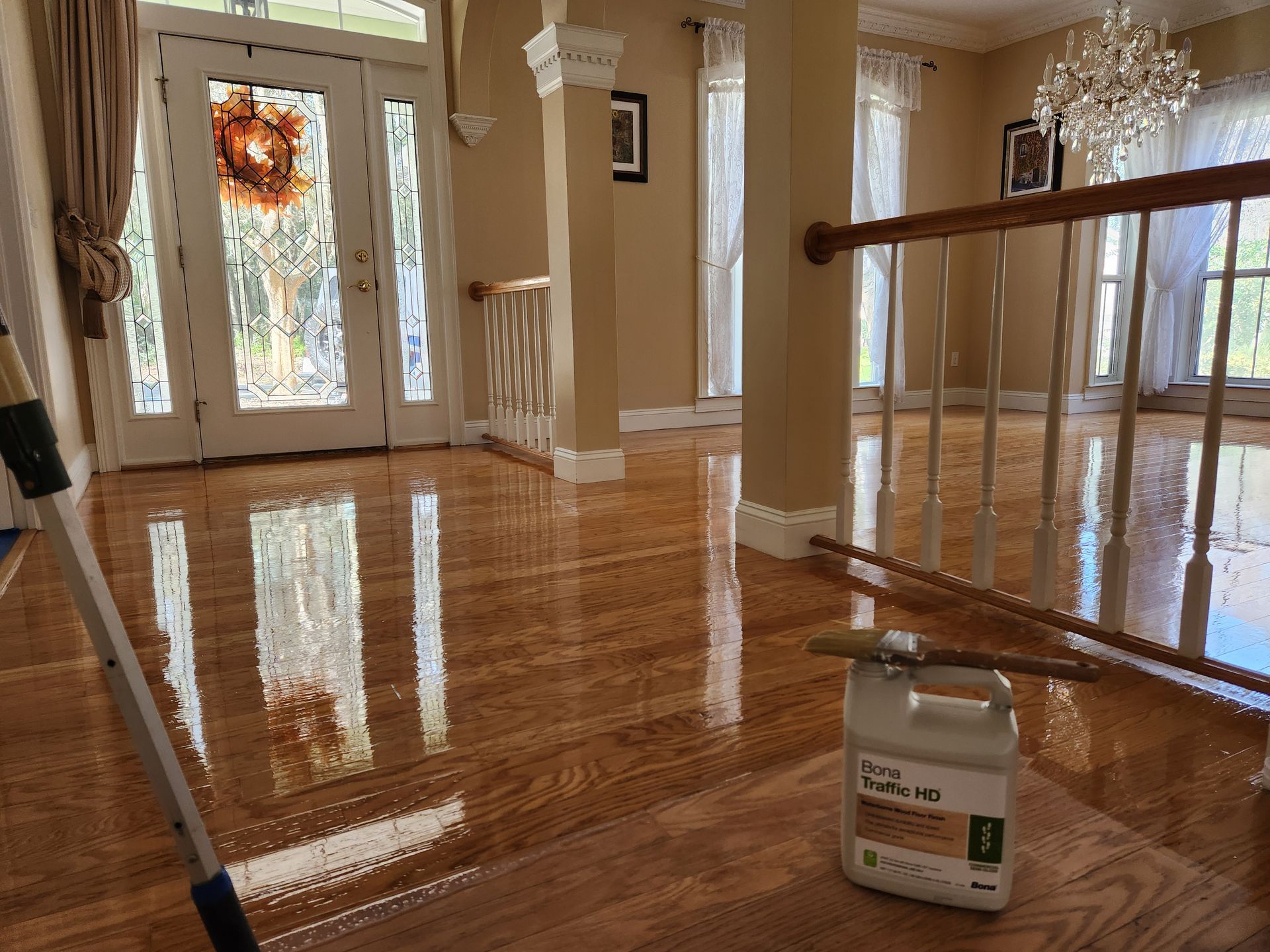 Freshly polished hardwood floor in a brightly lit foyer with a cleaning mop and a container of floor finish nearby.