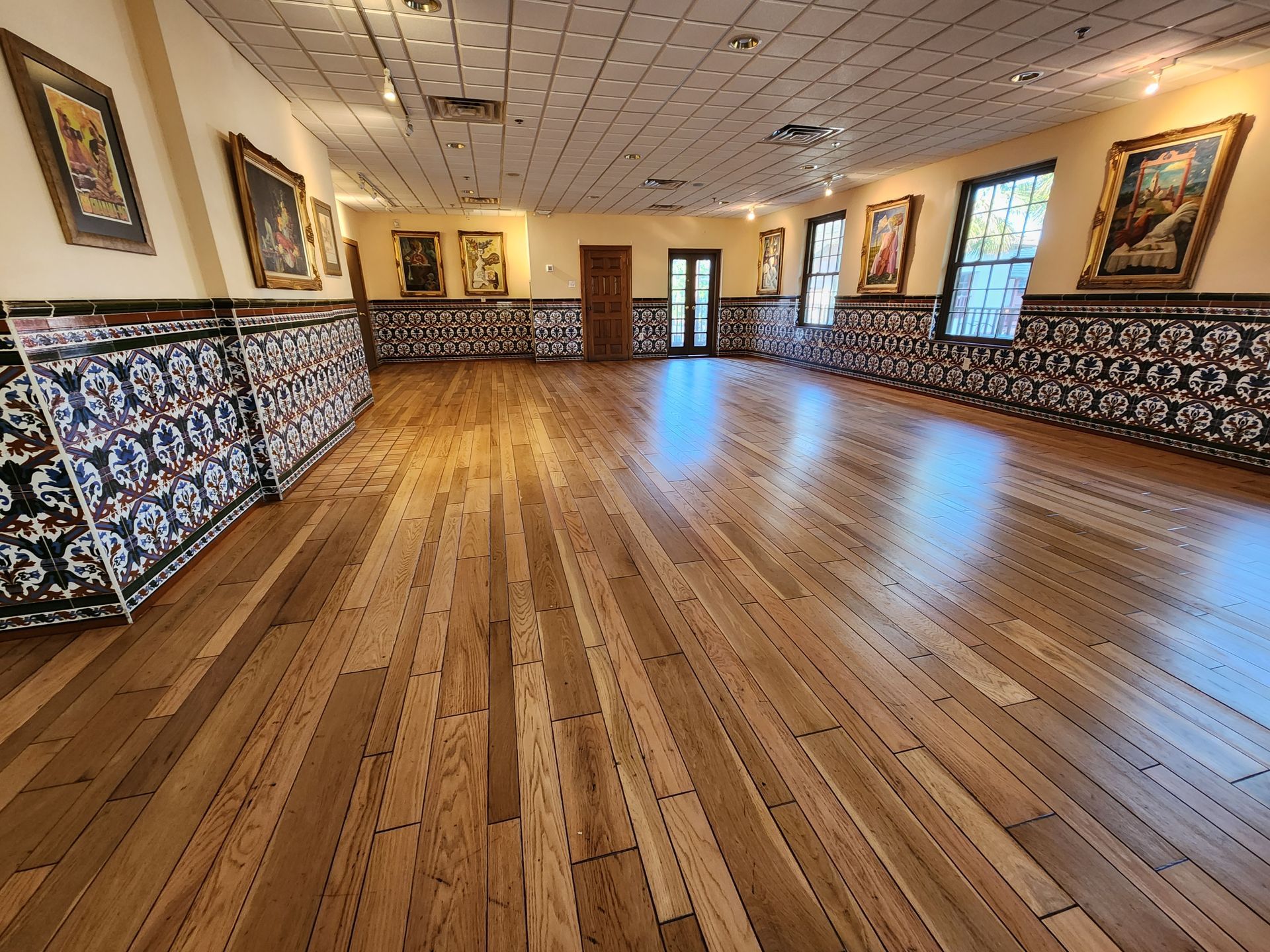 A spacious hall features wood floors, ornate tile wainscoting, and several framed paintings hanging on the pale walls.