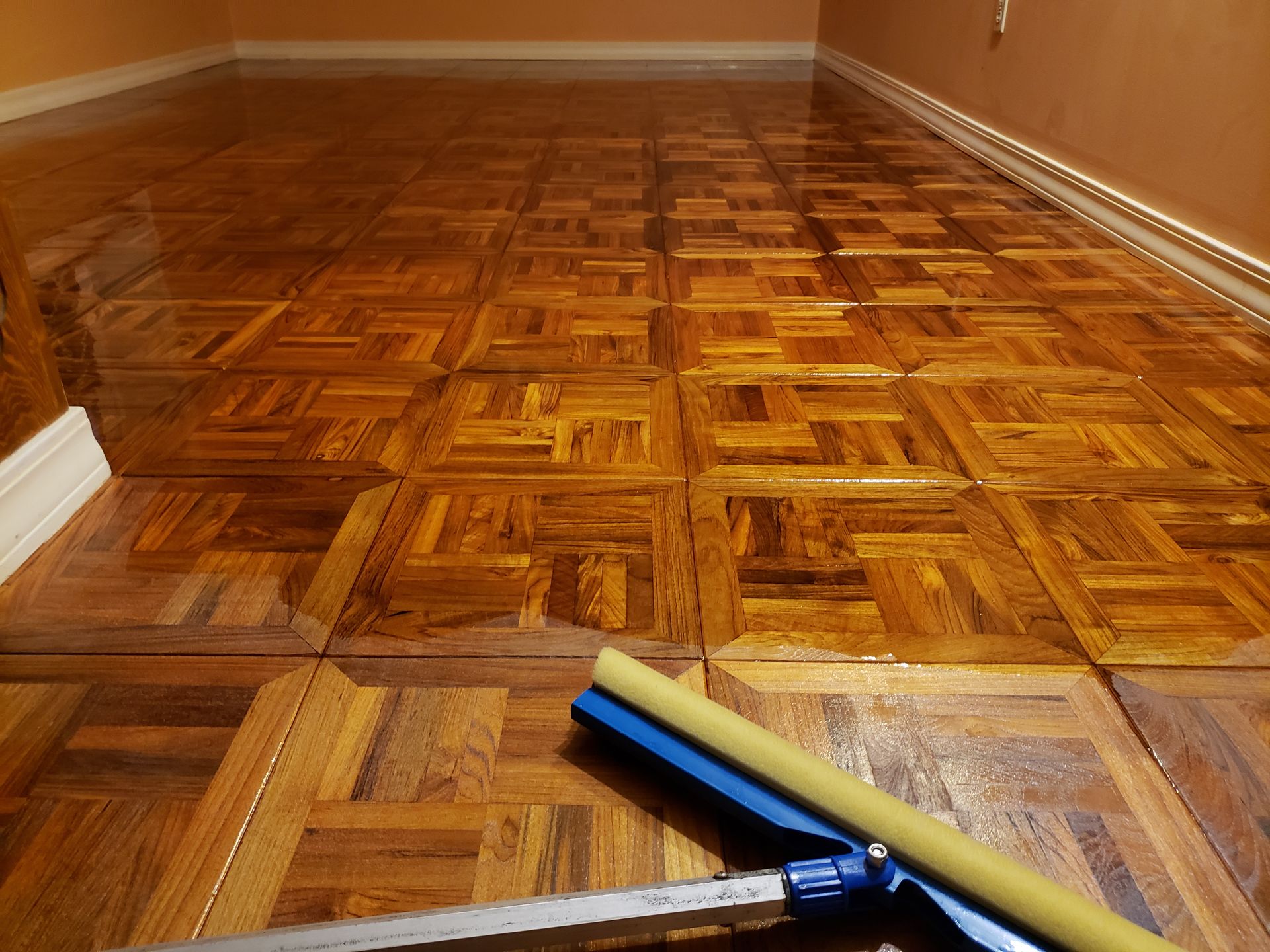A sponge mop rests on a freshly polished, shiny wooden parquet floor in a room with light brown walls.