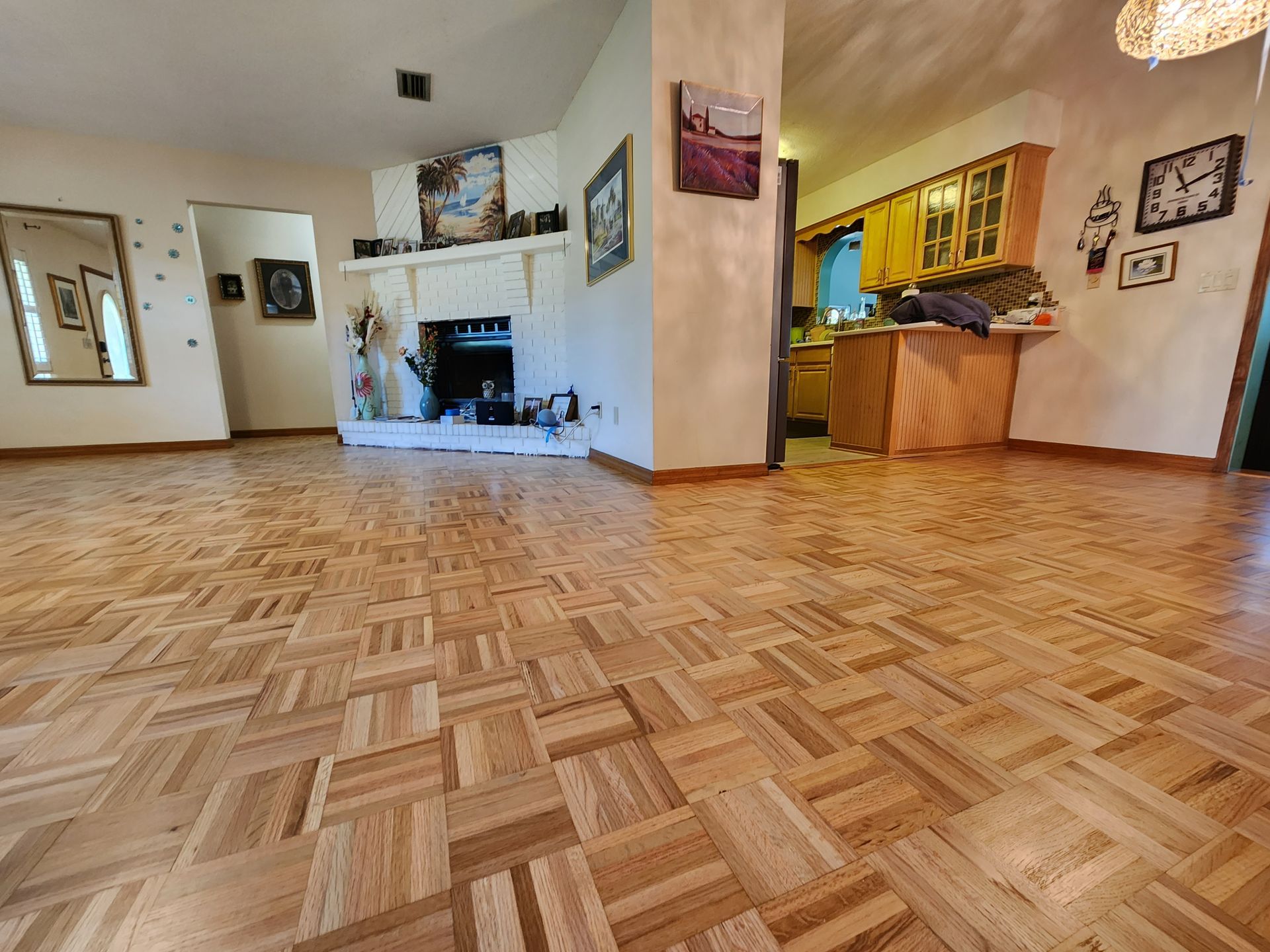 A wide-angle view of a bright living room with light-colored parquet flooring, a white fireplace, and an open kitchen.