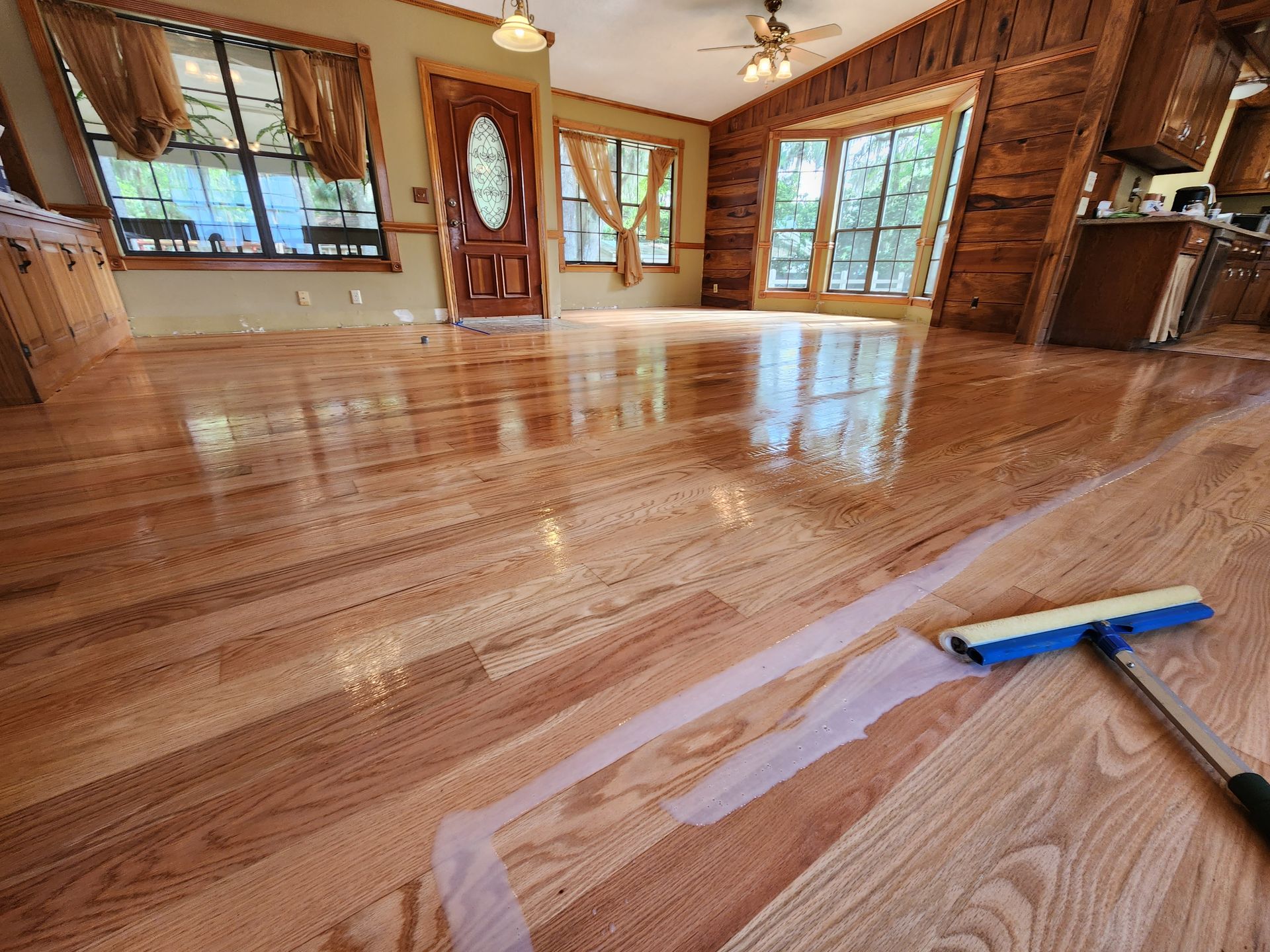 A squeegee on freshly refinished, glossy hardwood floors in a sunlit living room with wood-paneled walls.