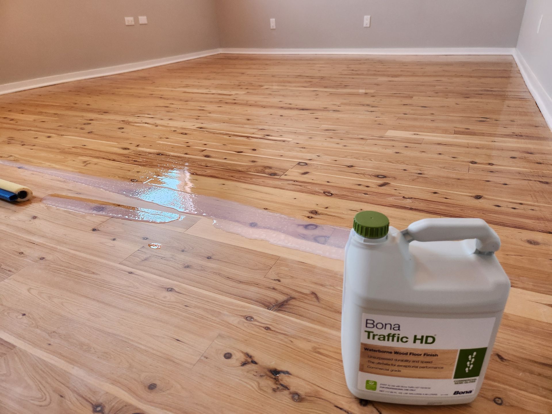 A jug of Bona Traffic HD floor finish rests on a wooden floor currently being coated with a finish applicator.