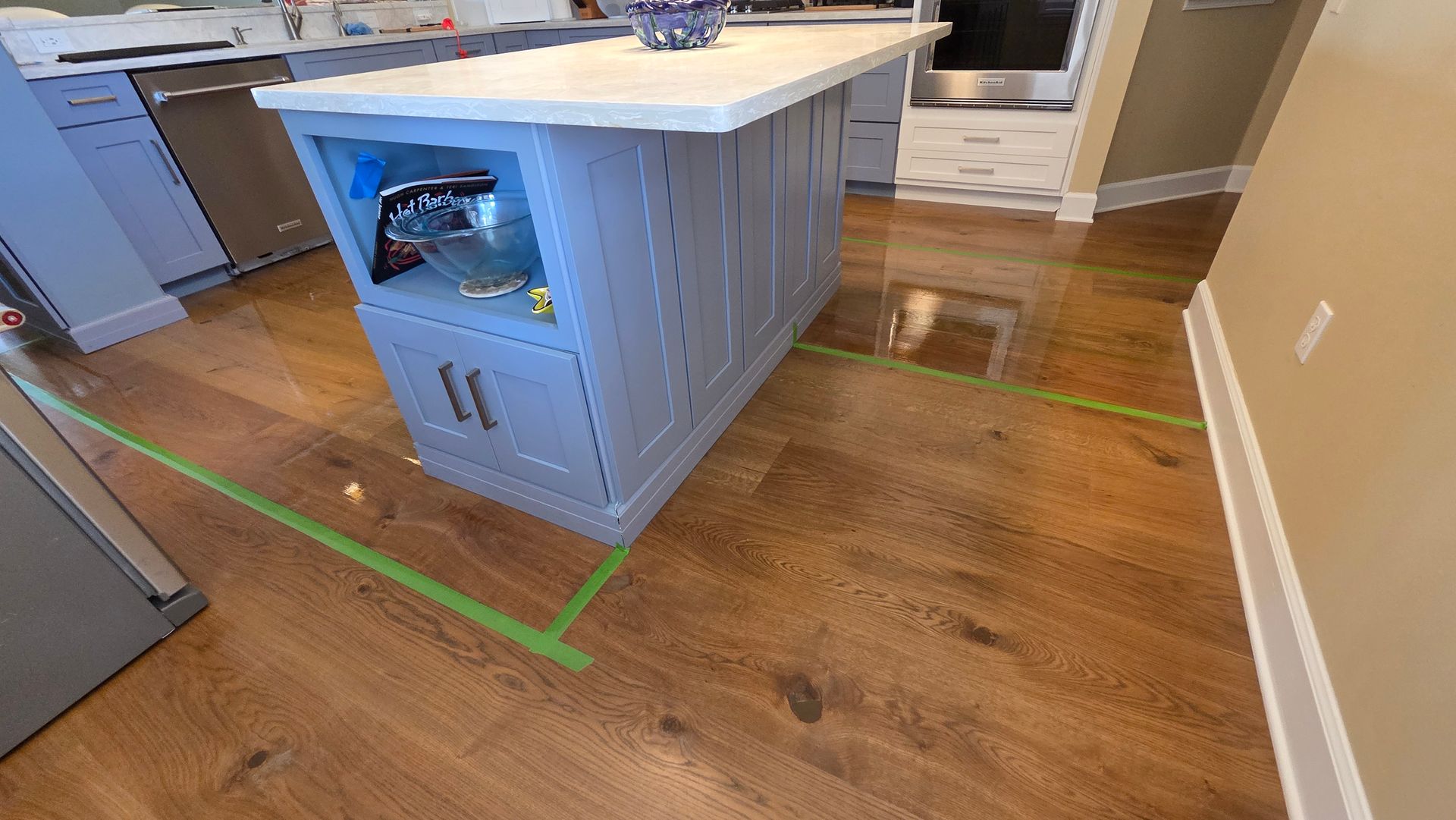 A kitchen with light-blue island cabinetry and hardwood floors, with green painter's tape outlining a work area.