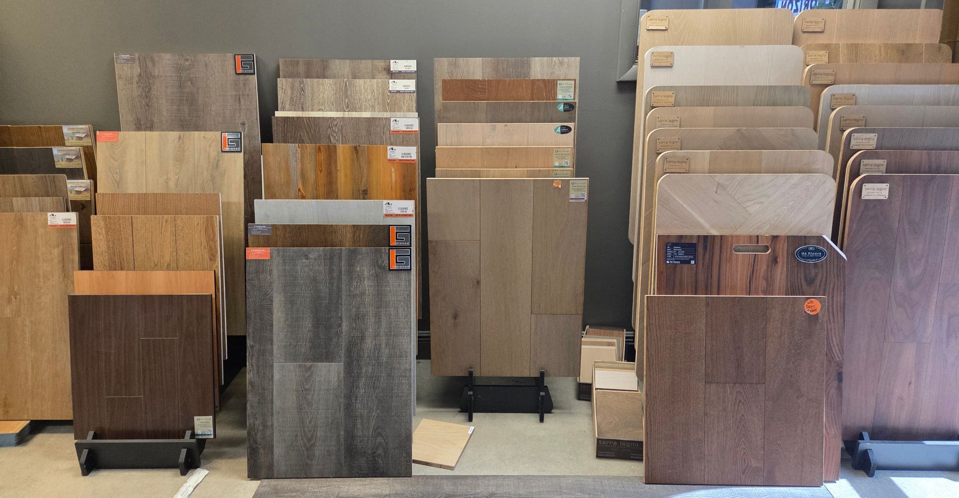 Store display showing multiple samples of wood and vinyl flooring planks in various shades of brown, gray, and beige.