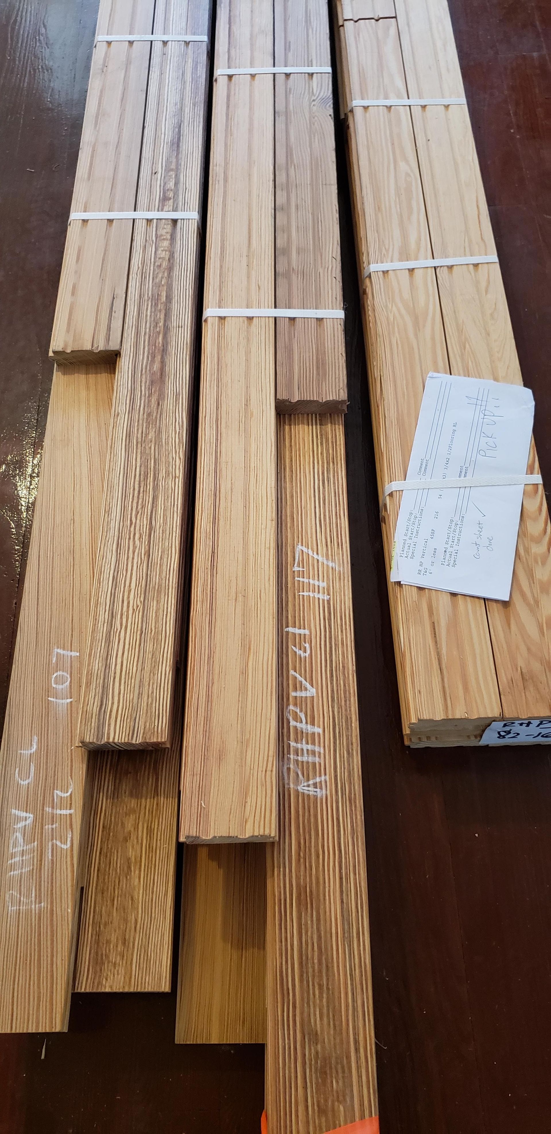 Three long, light-brown wood planks with distinct grain patterns arranged vertically on a dark surface, one with a paper tag.