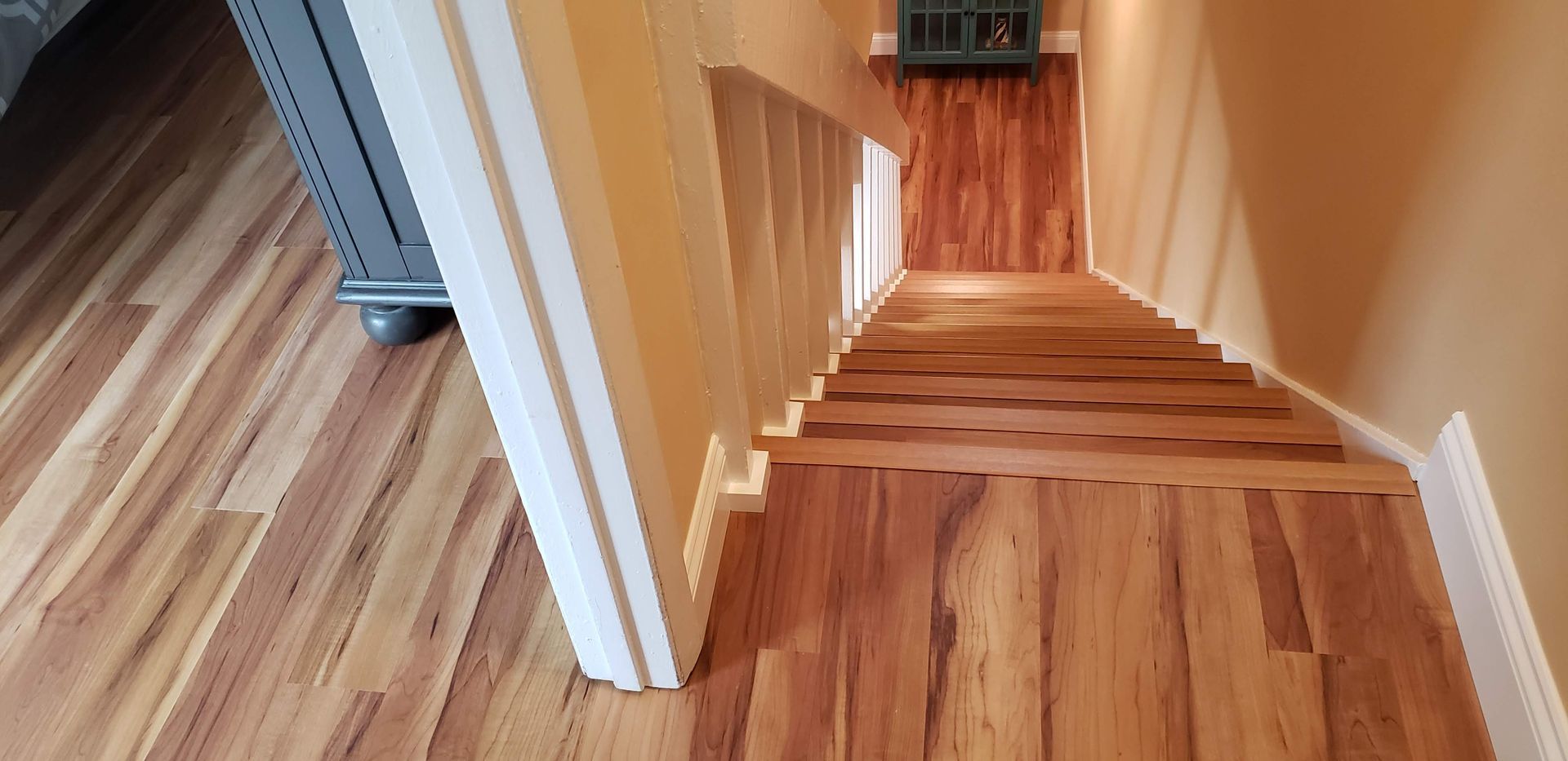 A staircase with wood flooring descends from a light-colored hallway, featuring white trim and a grey cabinet.
