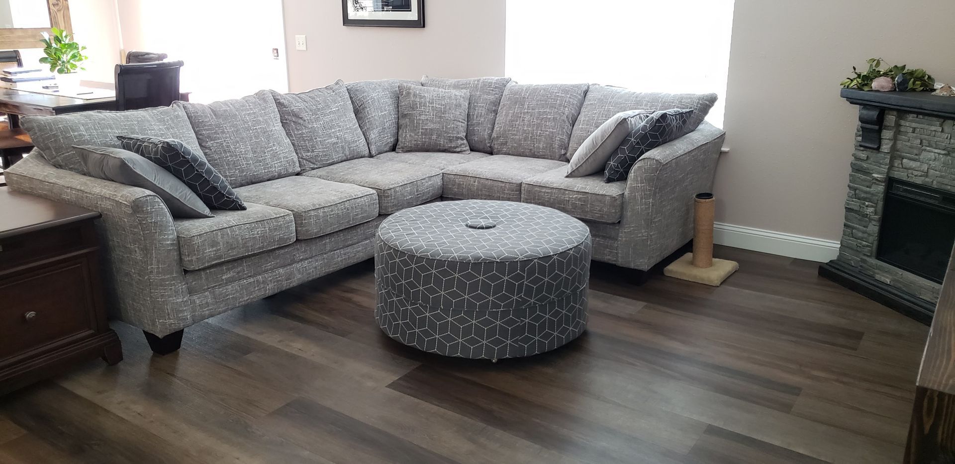 A gray patterned sectional sofa with a matching round ottoman on dark wood flooring in a room with a corner fireplace.