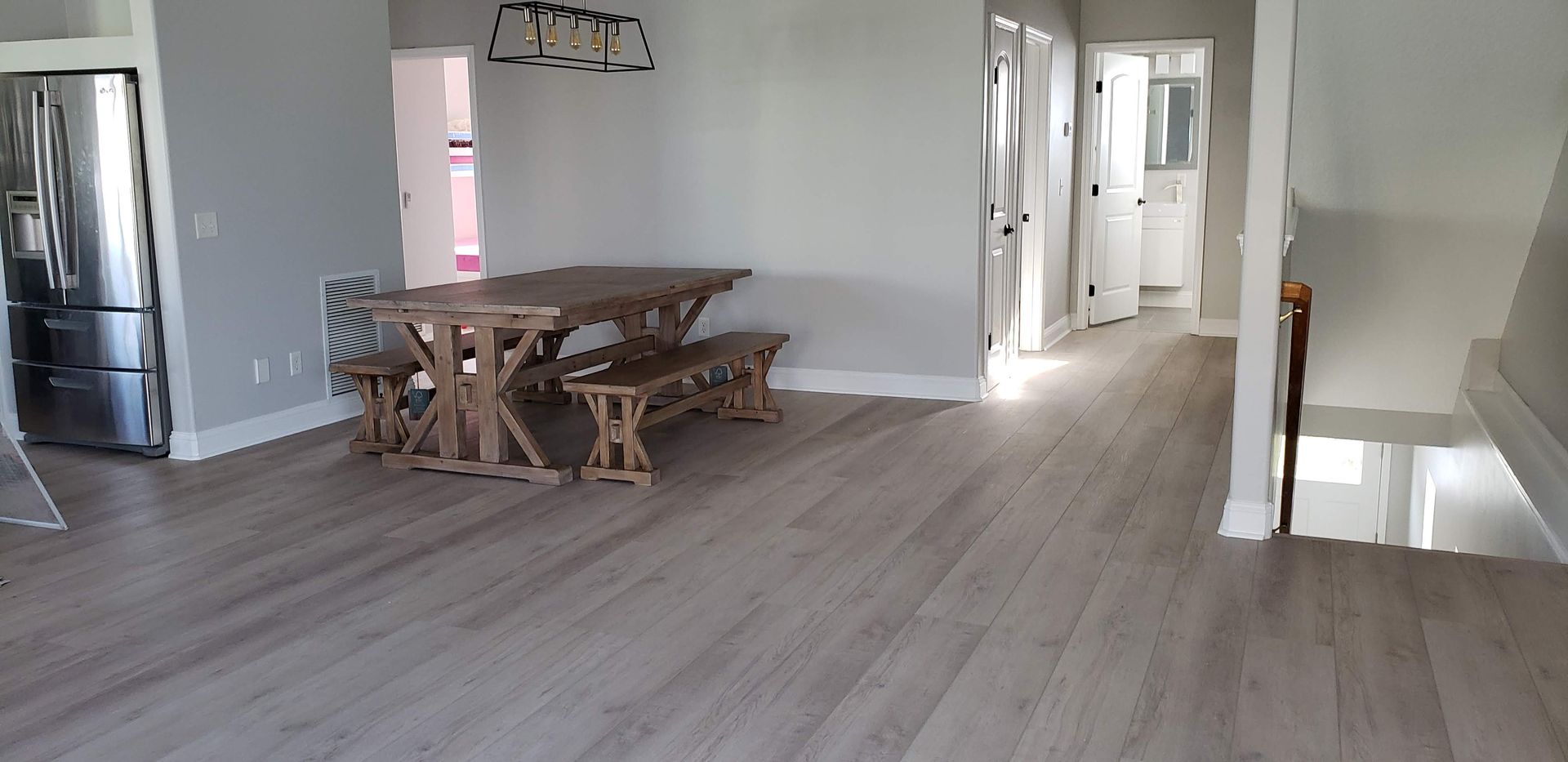 A dining area with a wooden table and two benches, light gray walls, and light wood floors, viewed from a large open room.